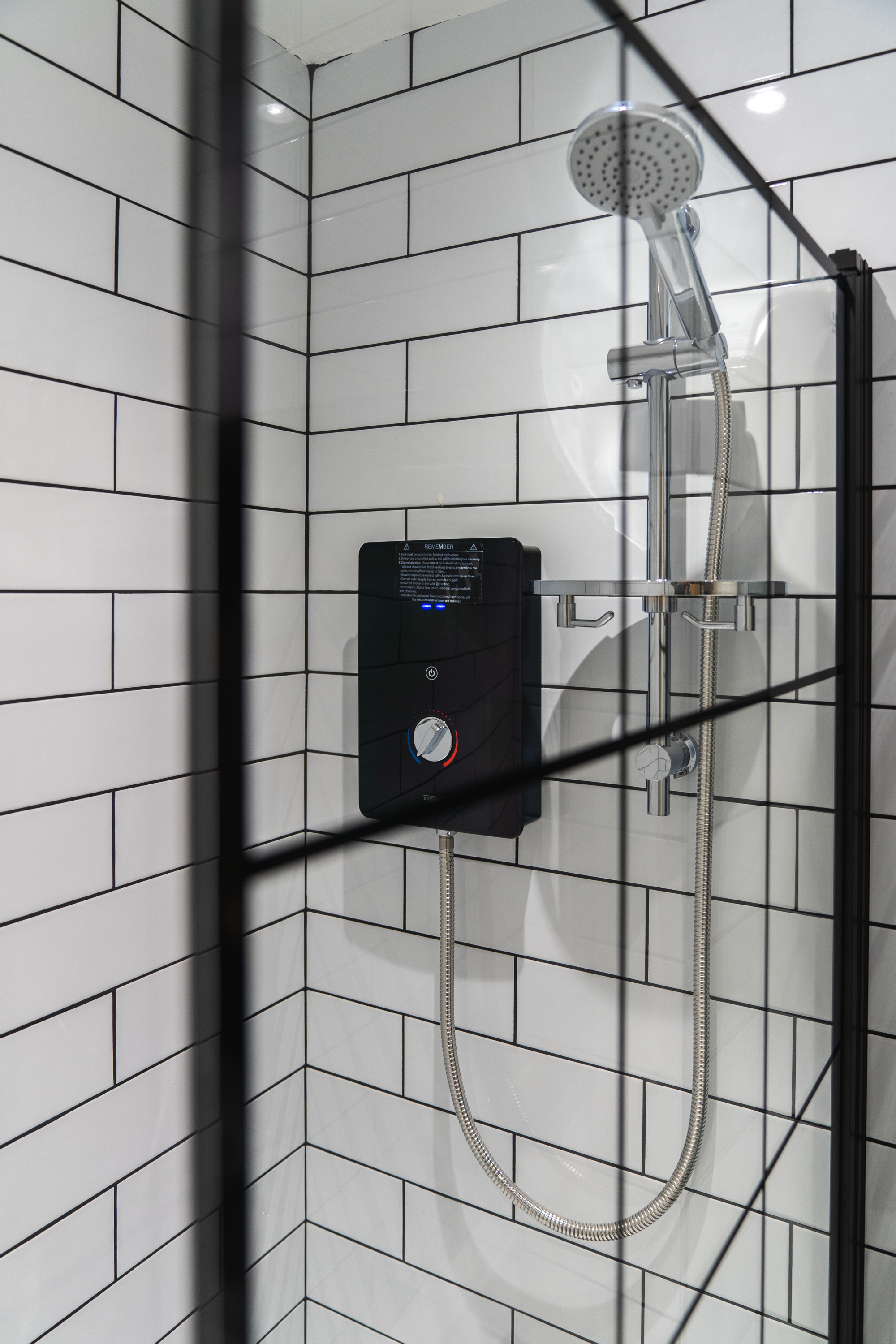 Shower with white subway tiles on the walls, a black electric shower heater with controls, a chrome showerhead, and a glass shower partition.