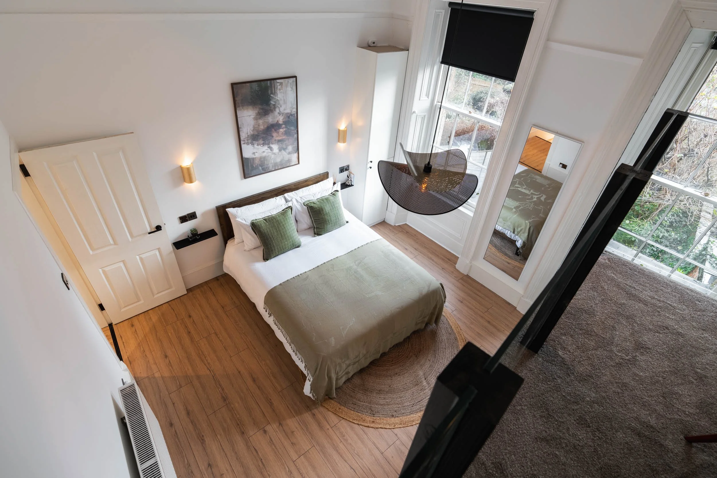 A spacious bedroom with a bed covered in a green blanket and matching green pillows, large windows with black curtains, a hanging light fixture, a floor mirror, and a wooden floor with a round rug.