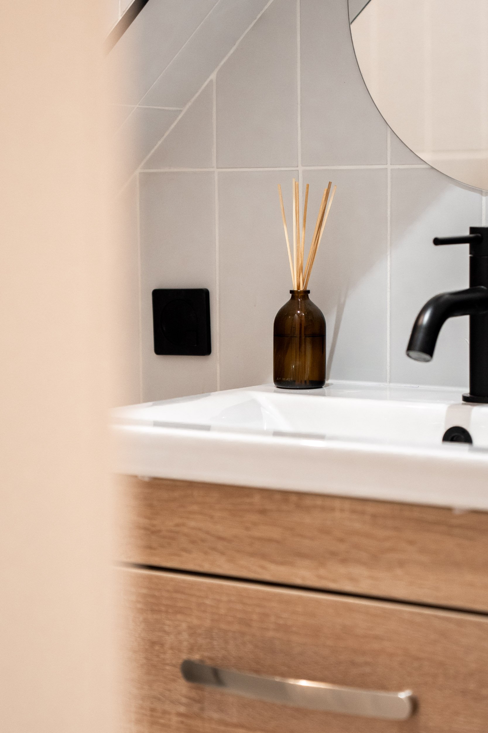 A bathroom sink area with a brown glass container holding reed diffuser sticks on the white countertop, black faucet, wooden cabinet, and tiled wall in the background.