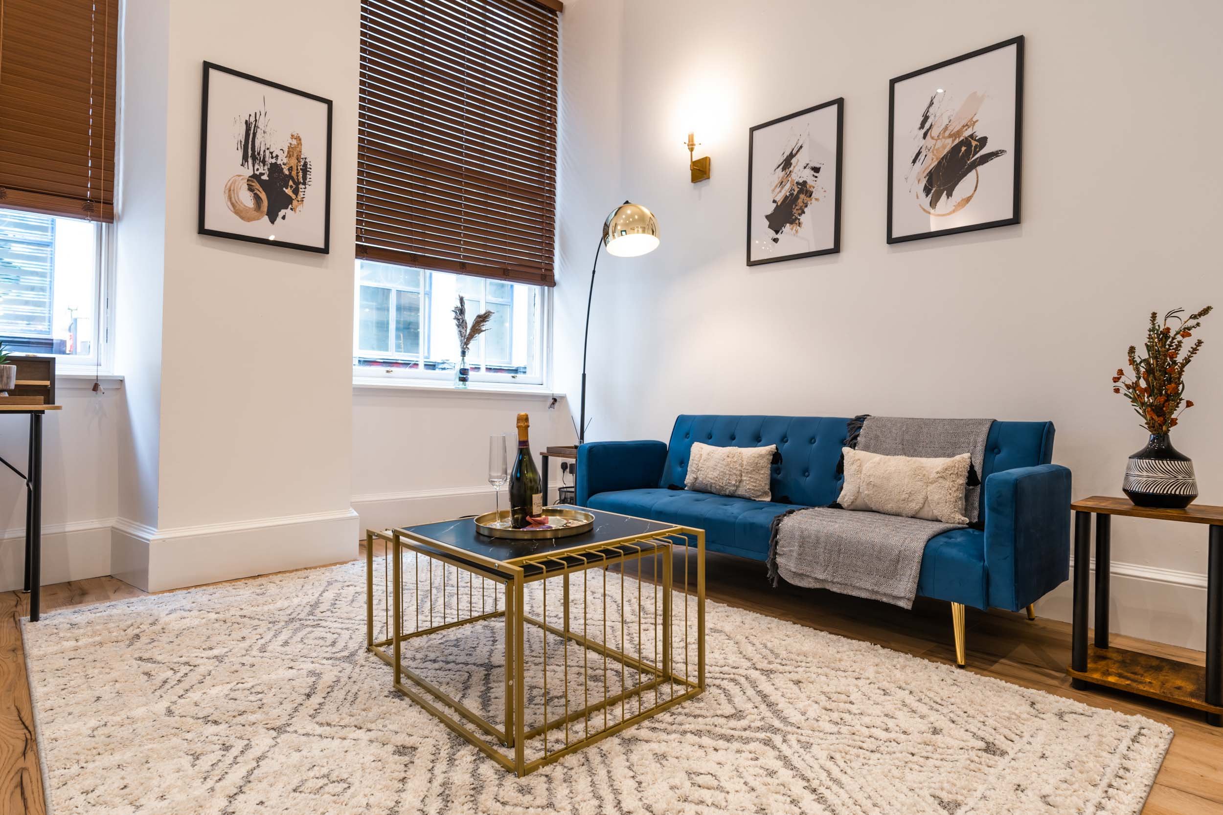 Living room with white walls, wooden floor, beige patterned rug, blue velvet sofa with pillows, black and gold side tables, gold framed coffee table with drinks, artwork on walls, window with brown blinds, lamp, and decorative vases.