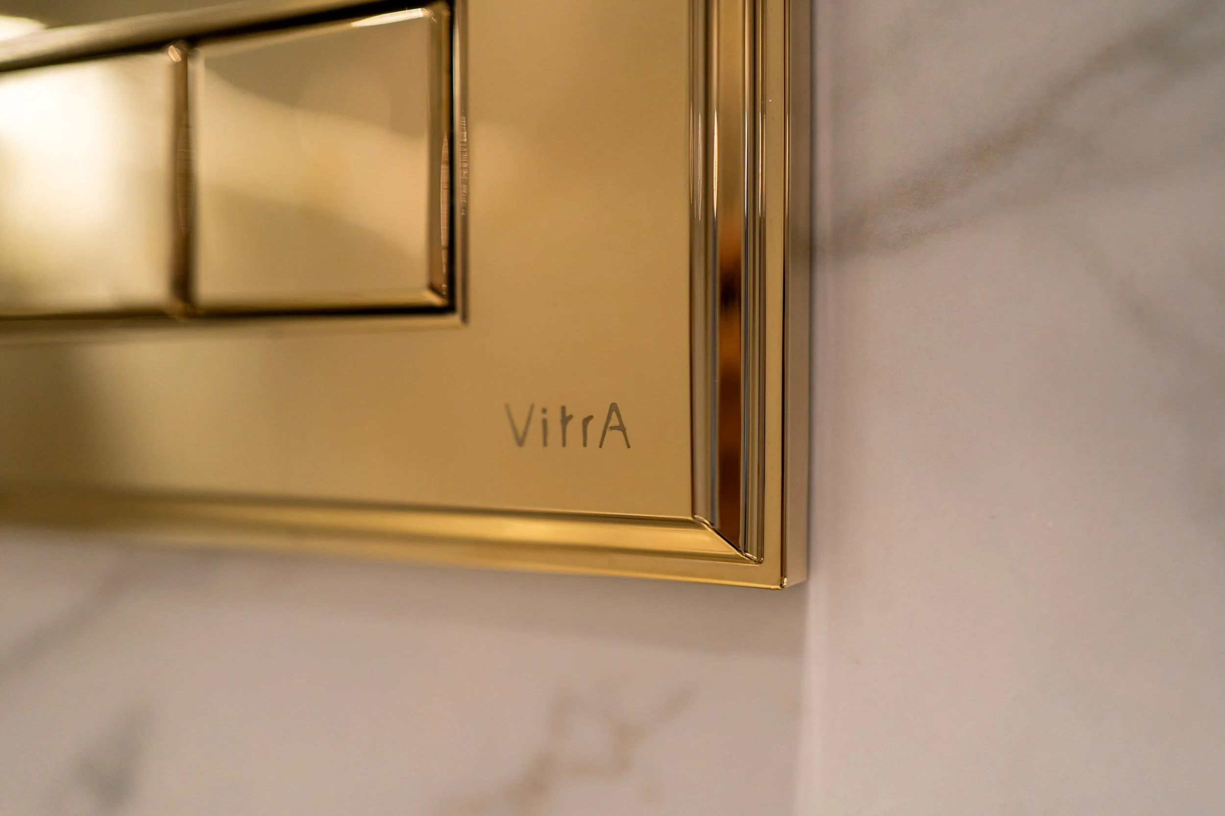 Close-up of a gold-colored flush button panel with the word 'Vitra' engraved on it, installed on a marble wall.