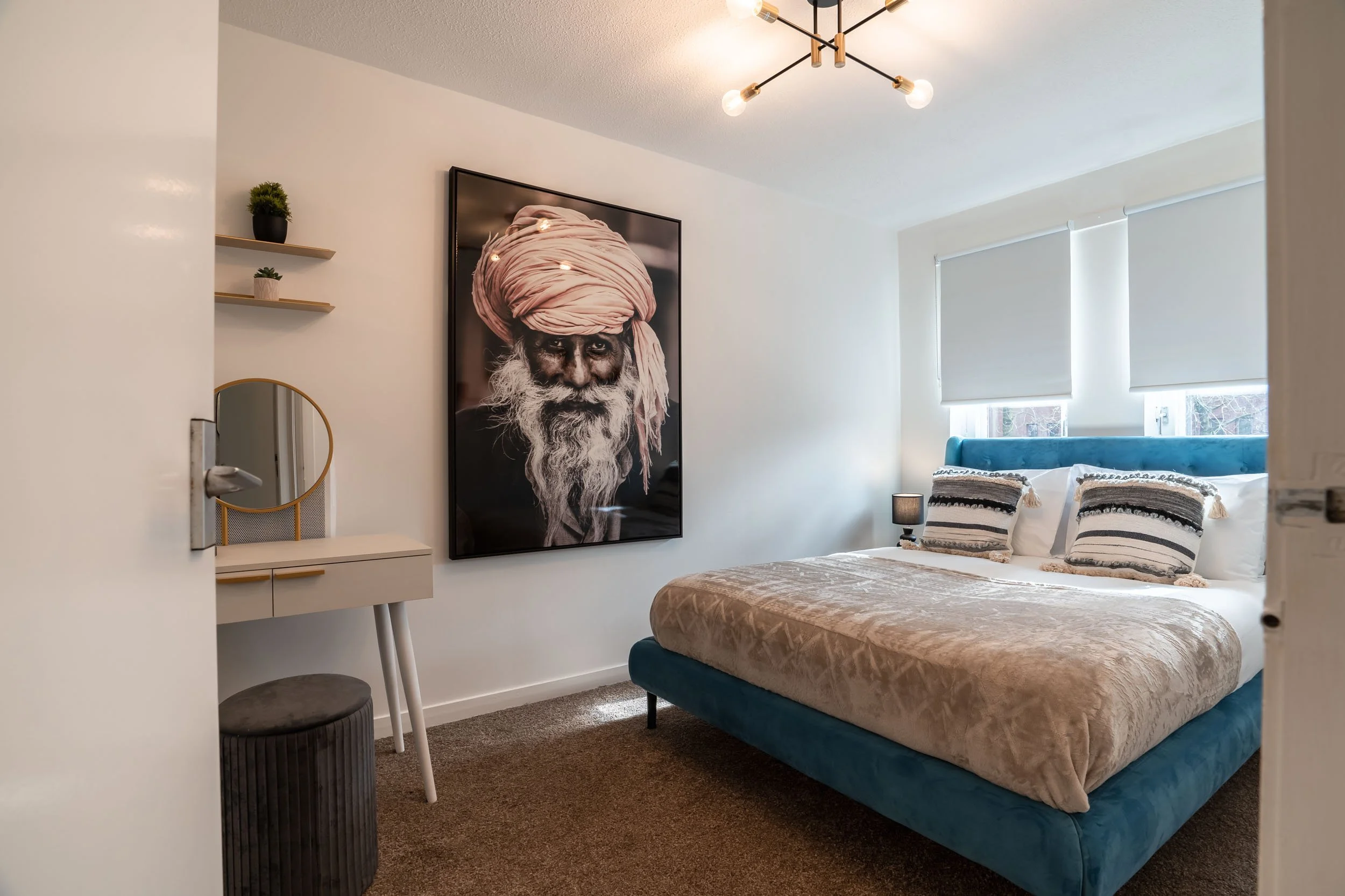 A cozy bedroom with a bed, two pillows with striped pillowcases, a side table with a lamp, a large portrait of an elderly man with a turban on the wall, two small wall-mounted shelves with potted plants, and a modern ceiling light fixture.