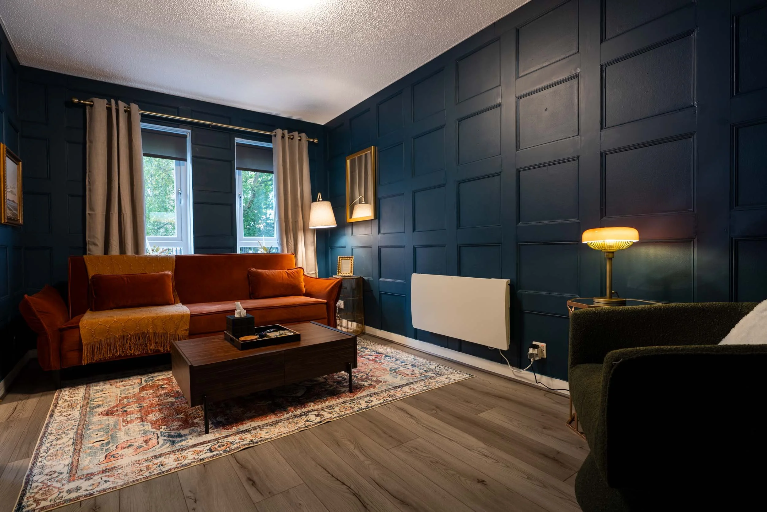 Living room with dark blue paneled walls, beige curtains, a rust-colored sofa, a green armchair, a wooden coffee table, a patterned rug, a table lamp, a wall mirror, and a window showing greenery outside.