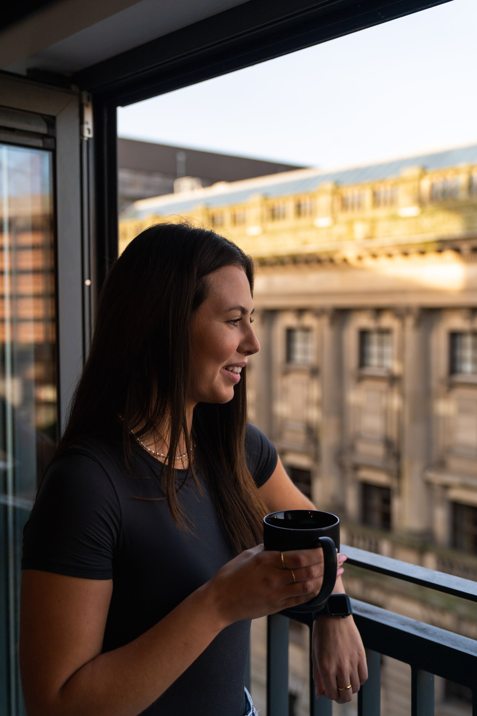 A woman with long dark hair smiling while holding a black coffee mug on a balcony, with a cityscape background of buildings and an elevated train passing by.