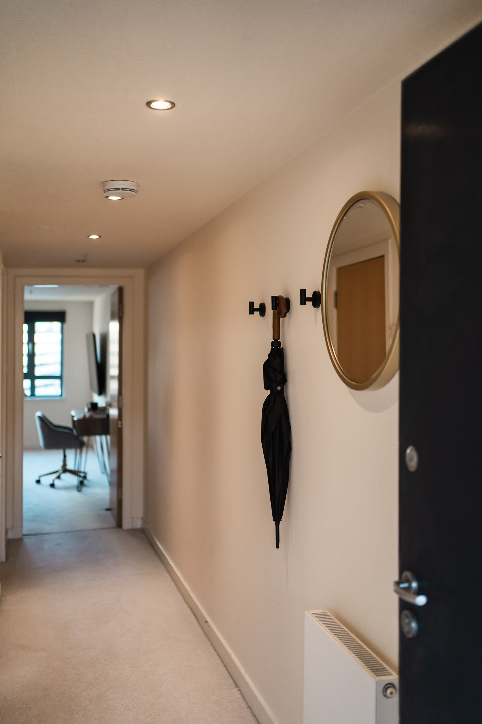 A hallway with a ceiling with built-in lights, a smoke detector, and a wall-mounted coat hook holding a black umbrella and a wooden walking stick. A round mirror with a gold frame is on the wall, and a black doorway partially opens to reveal a room w