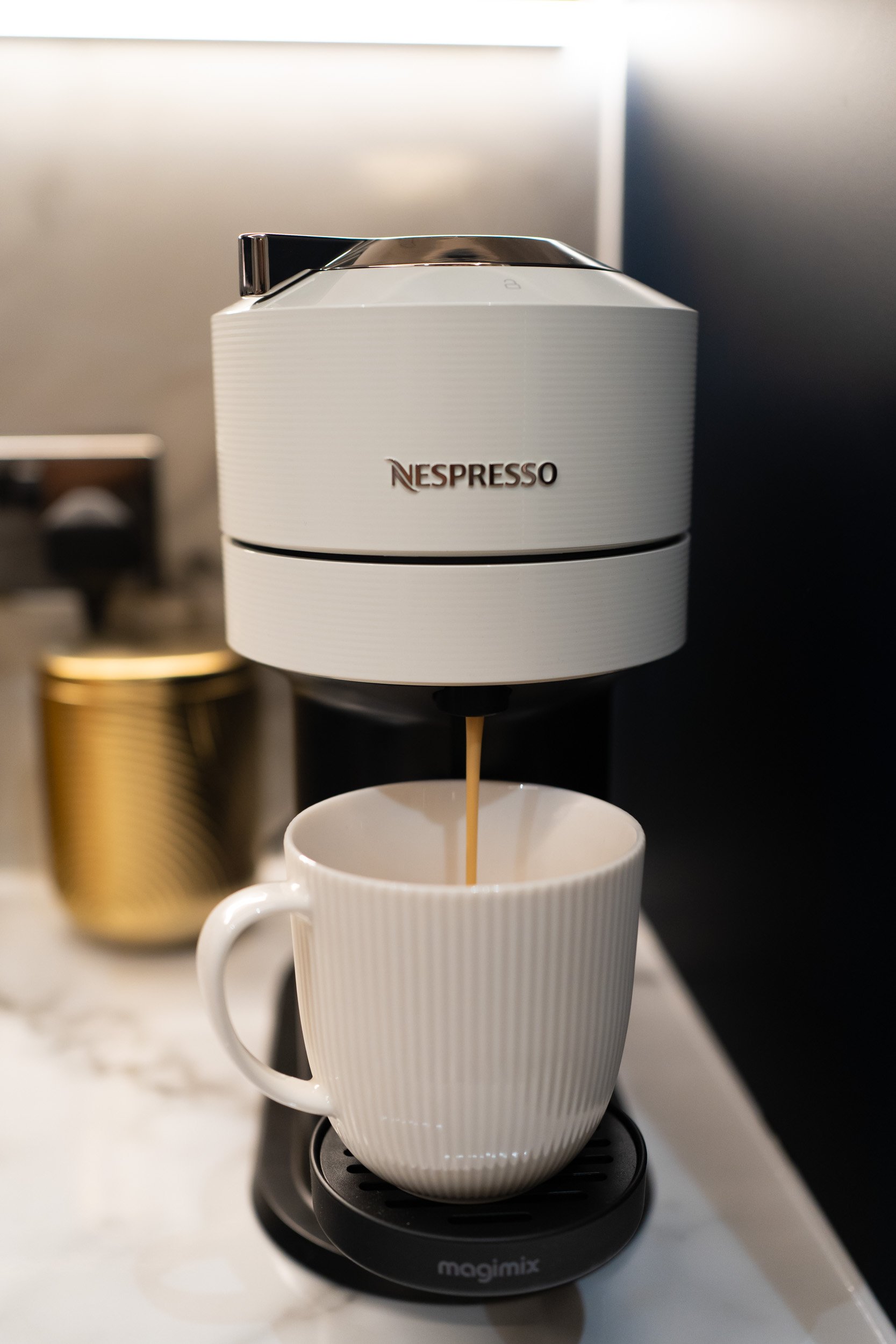 A Nespresso coffee machine is pouring coffee into a white cup placed on a Magimix stand, with a blurred background.