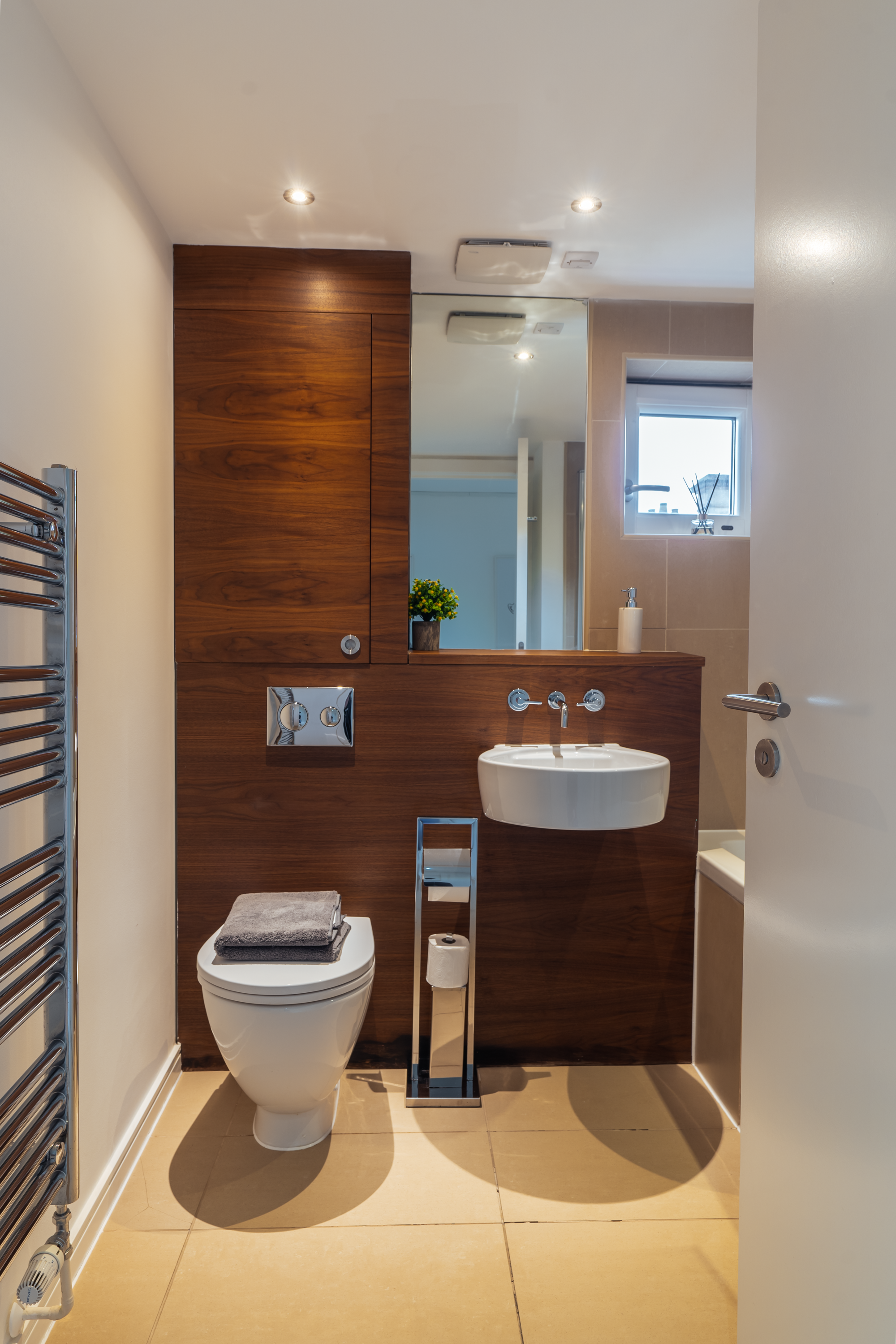 Modern bathroom with a white toilet with gray towels on it, a small white sink, a wooden wall with a mirror, a small window with a reed diffuser, a towel heater on the left, and a toilet paper holder in the center.