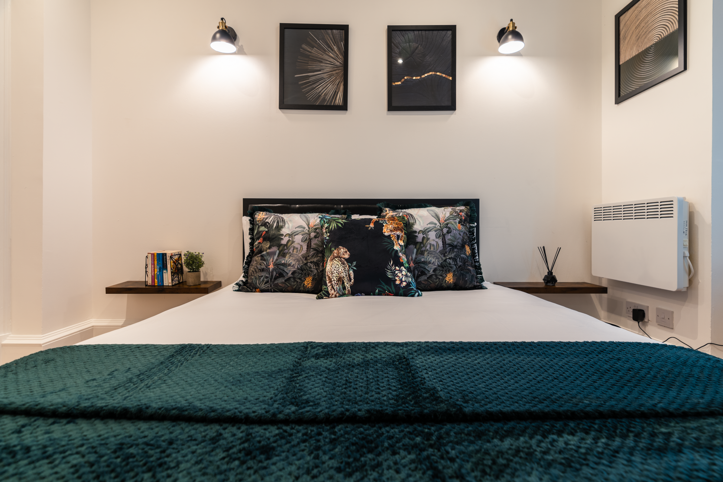 A neatly made bed with tropical-themed pillows and a dark green textured blanket at the foot. Two floating wooden nightstands on each side hold books, a small plant, a reed diffuser, and an unplugged electronic device. Above the bed, three framed abs