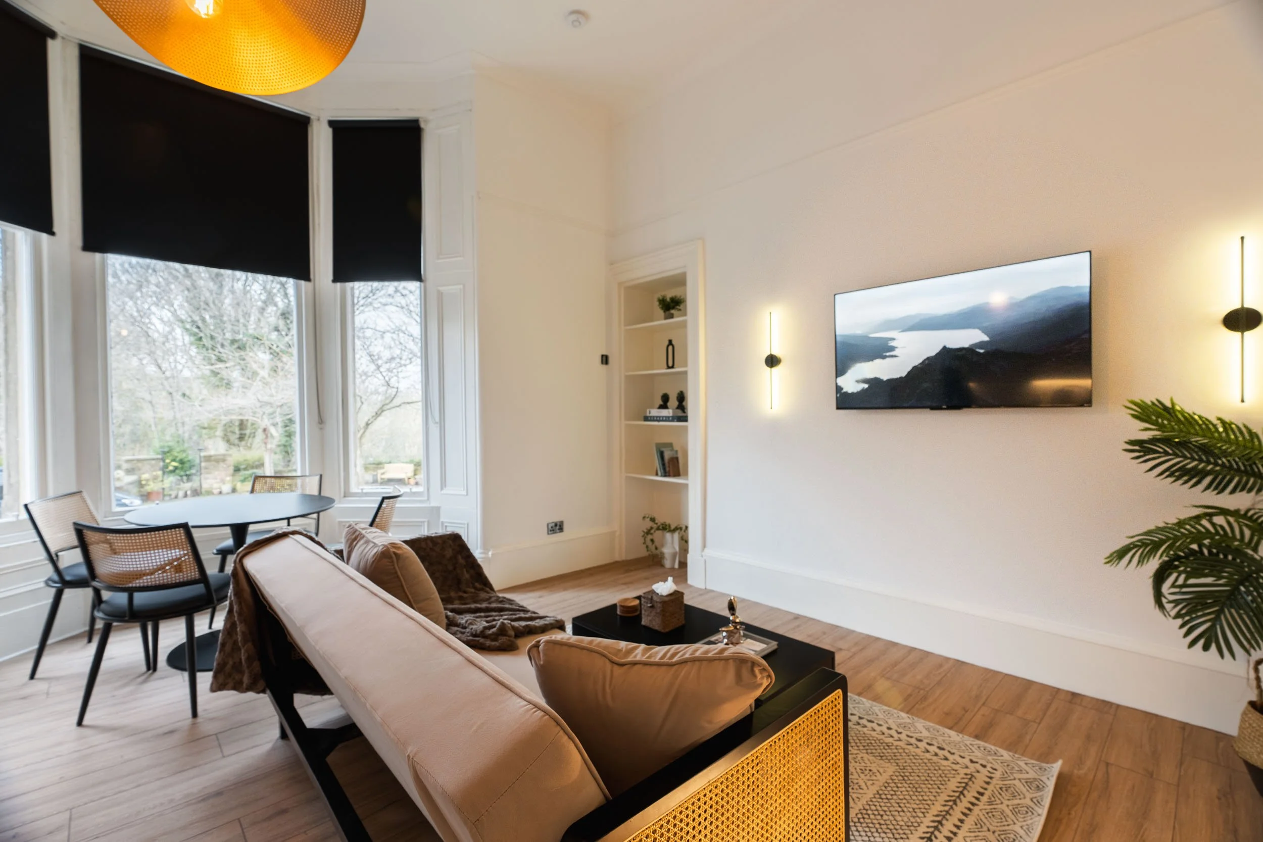 Living room with beige sofa, black coffee table, and a yarmulke on the table. Wall-mounted TV showing a landscape view. White built-in shelving with decor. Large windows with black blinds, hardwood floor, and potted plant in the corner.