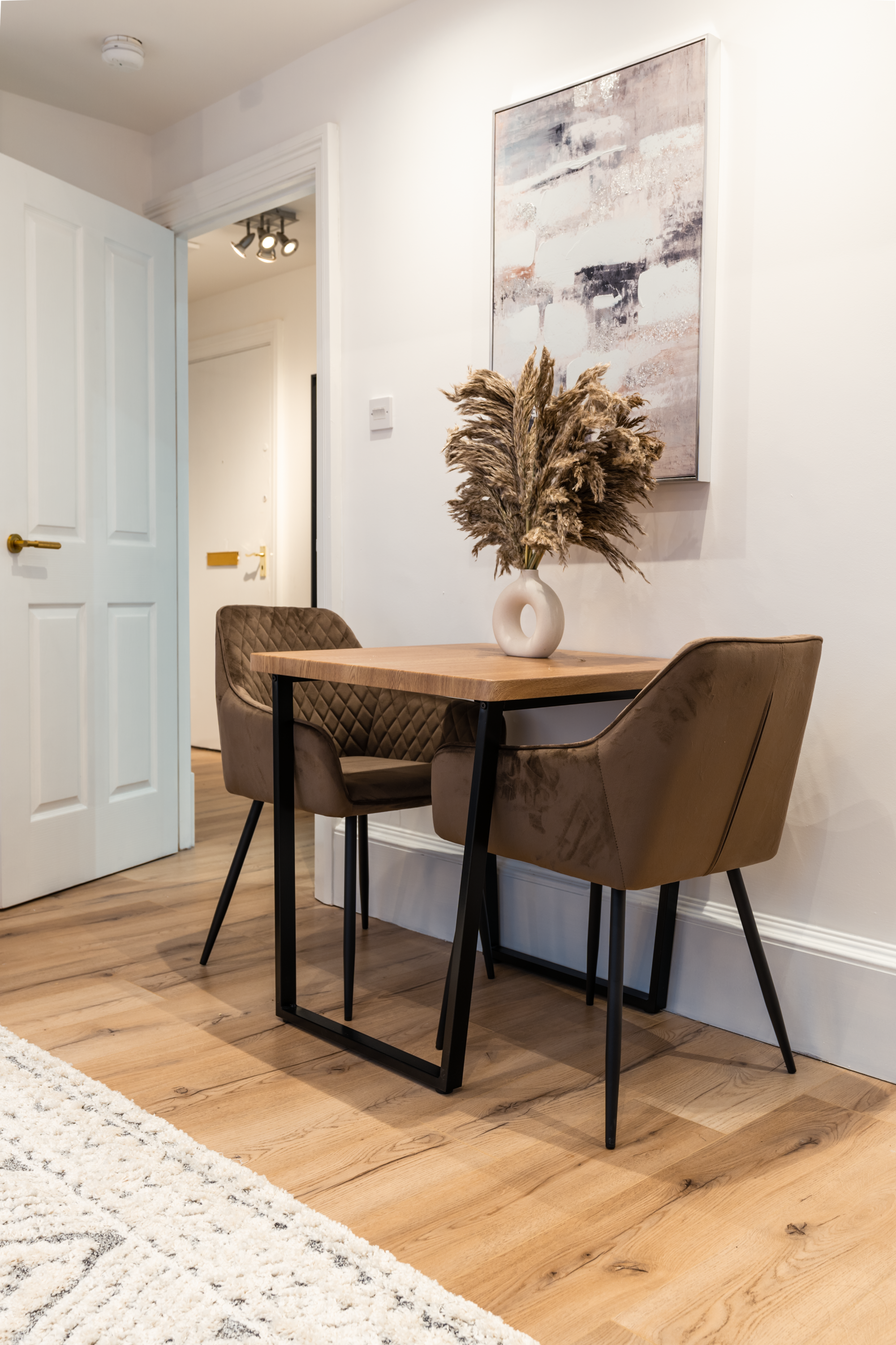 A small wooden dining table with a palette of beige and brown seed in a white-walled room. Two brown upholstered chairs with black metal legs are positioned on each side of the table. A white ceramic vase with dried pampas grass is on the table. A la