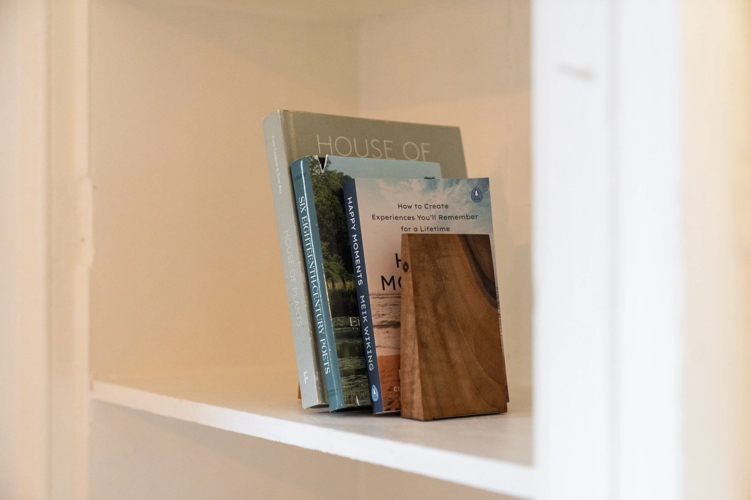 Four books standing upright on a white shelf, with one leaning against the others. The books are about homes, poetry, and experiences, with titles including 'HOUSE OF PLANTS,' 'SIX EIGHTEENTH CENTURY POETS,' 'HAPPY MOMENTS,' and a guide on creating m