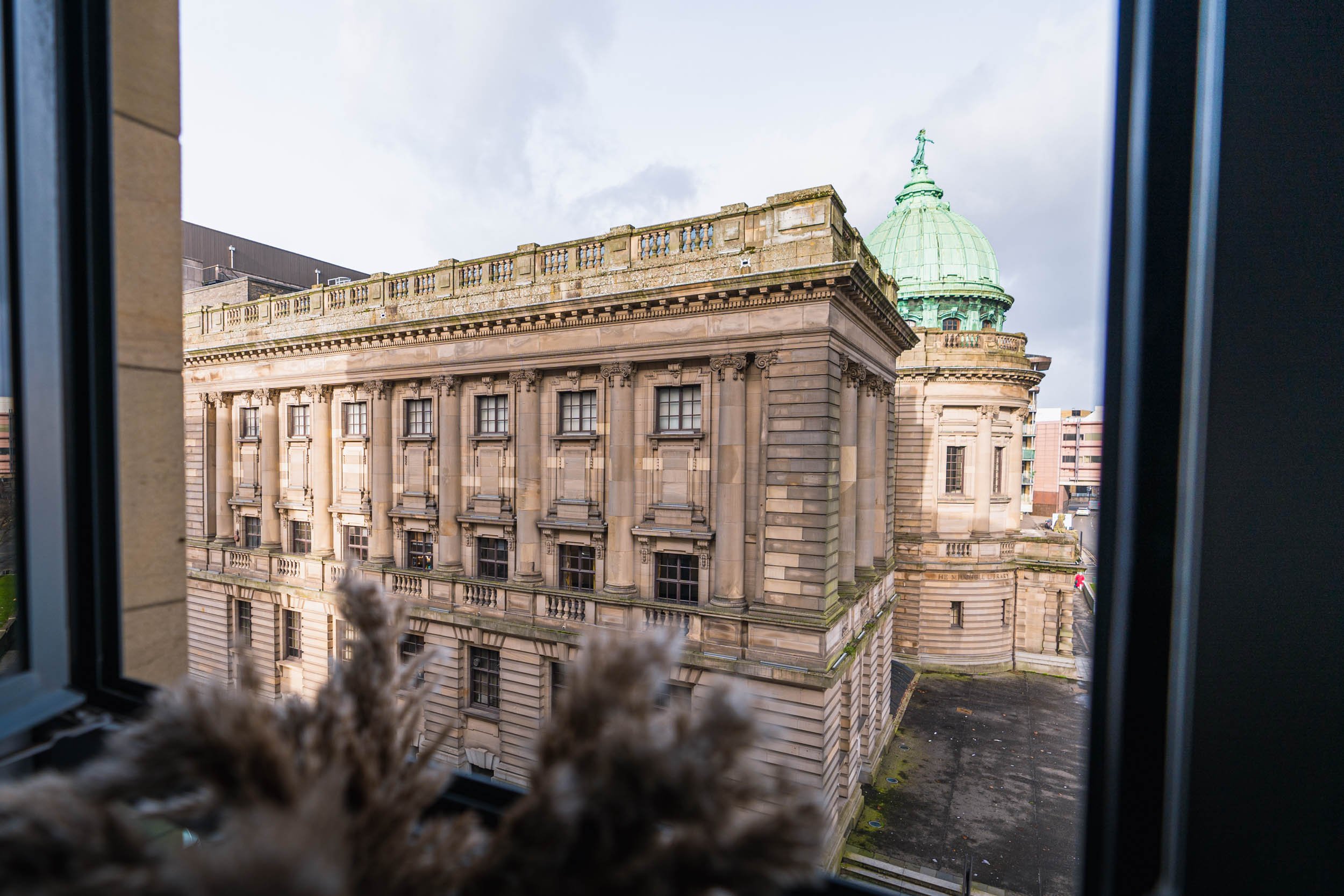 View of a historic building with detailed stone architecture and a green domed roof seen through a window with blue frames. Some plants are visible in the foreground.