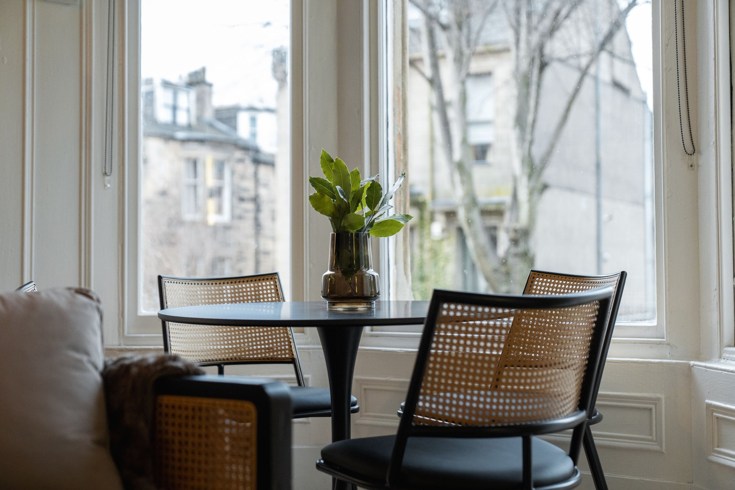 A round black table with a green vase containing leaves, placed near large windows with a view of trees and buildings outside.