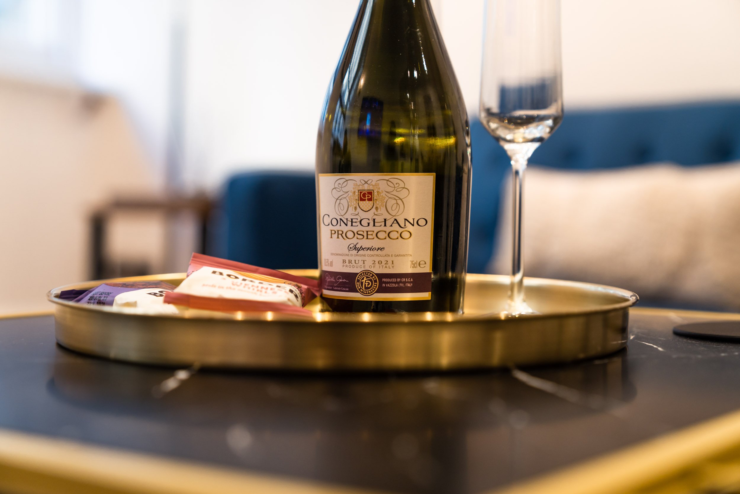 A tray holding a bottle of Conegliano Prosecco and a flute glass, along with some snack packets, on a dark table with a blurred background.