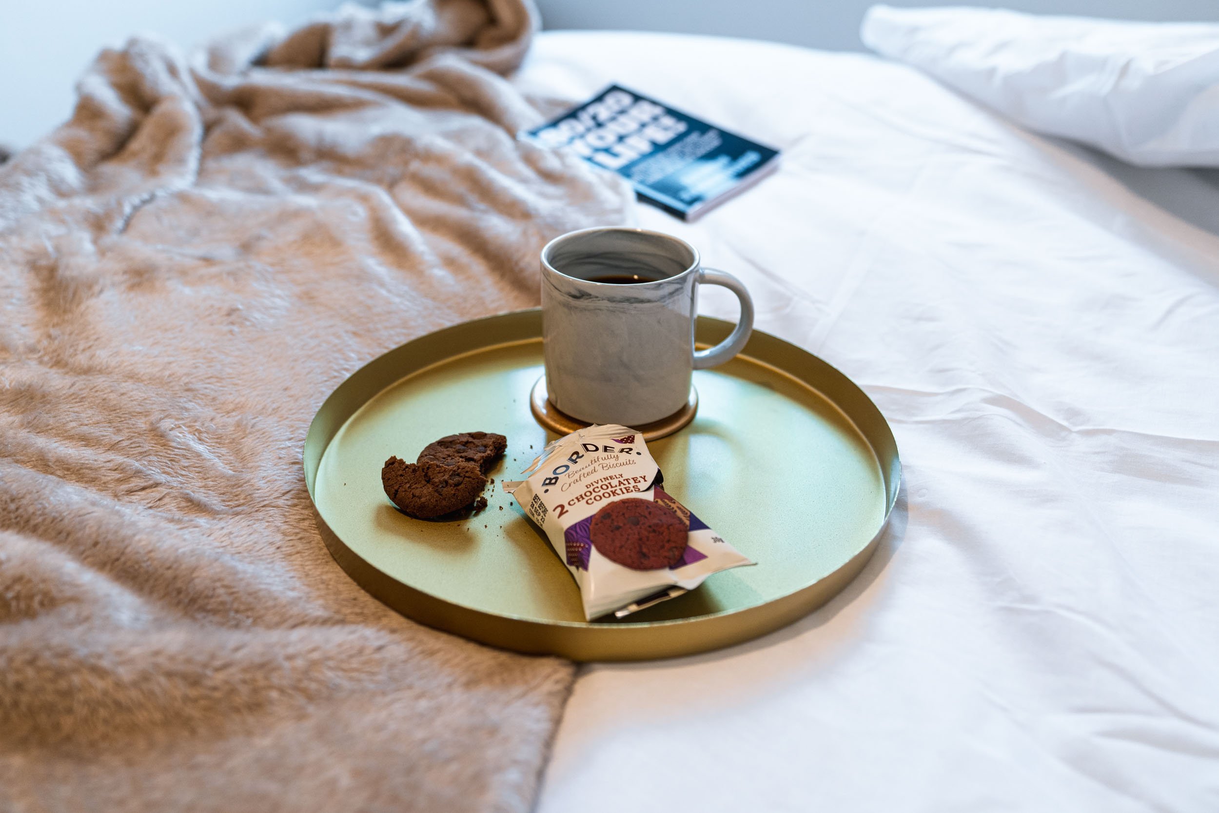 A breakfast tray with a mug of coffee, a partially eaten chocolate cookie, and a packaged chocolate chip cookie on a green round tray placed on a bed with beige and white bedding, a fuzzy beige blanket, and a magazine in the background.