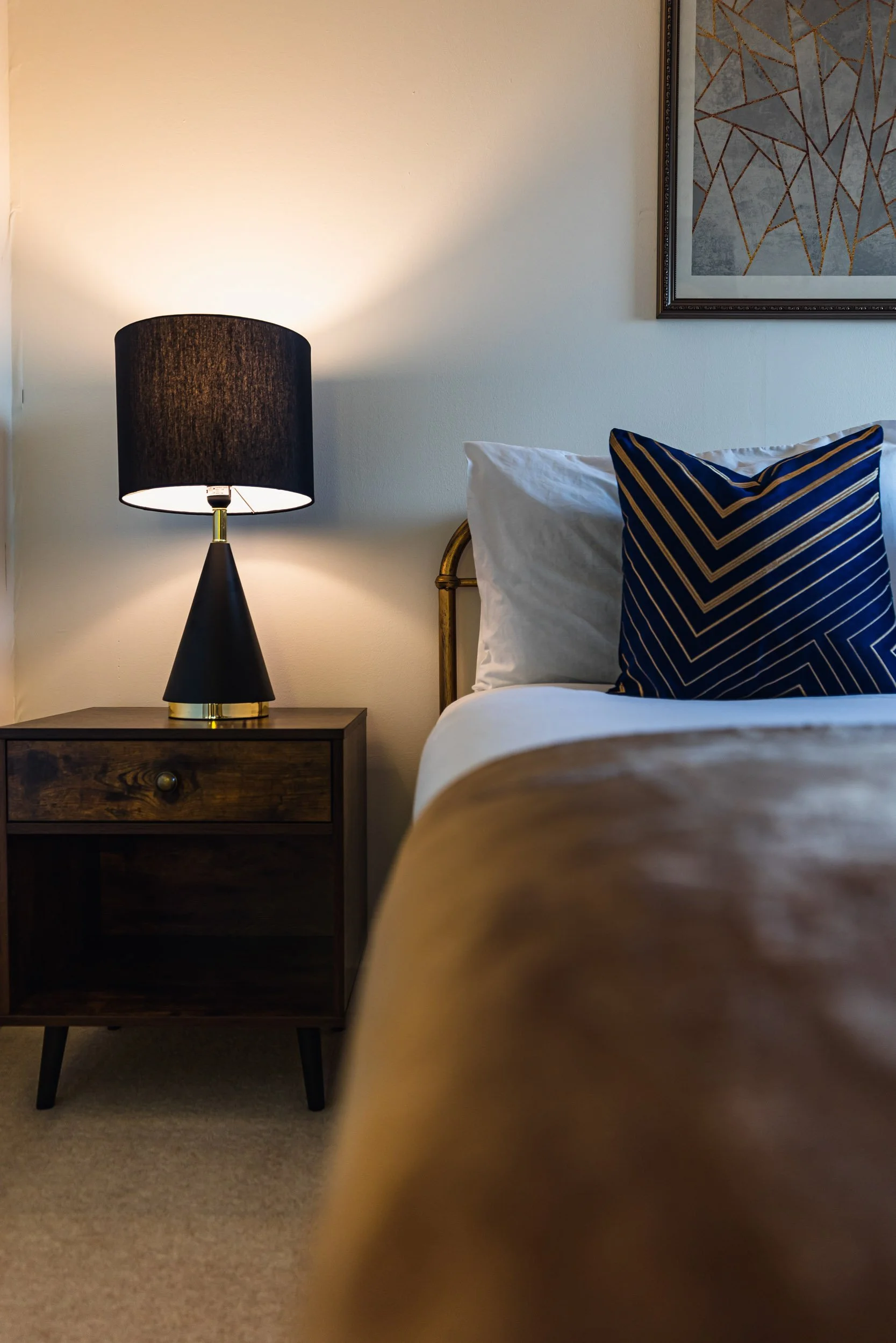 A bedroom corner with a wooden nightstand, black lampshade lamp, bed with white pillows and a navy blue decorative pillow, framed artwork on the wall.