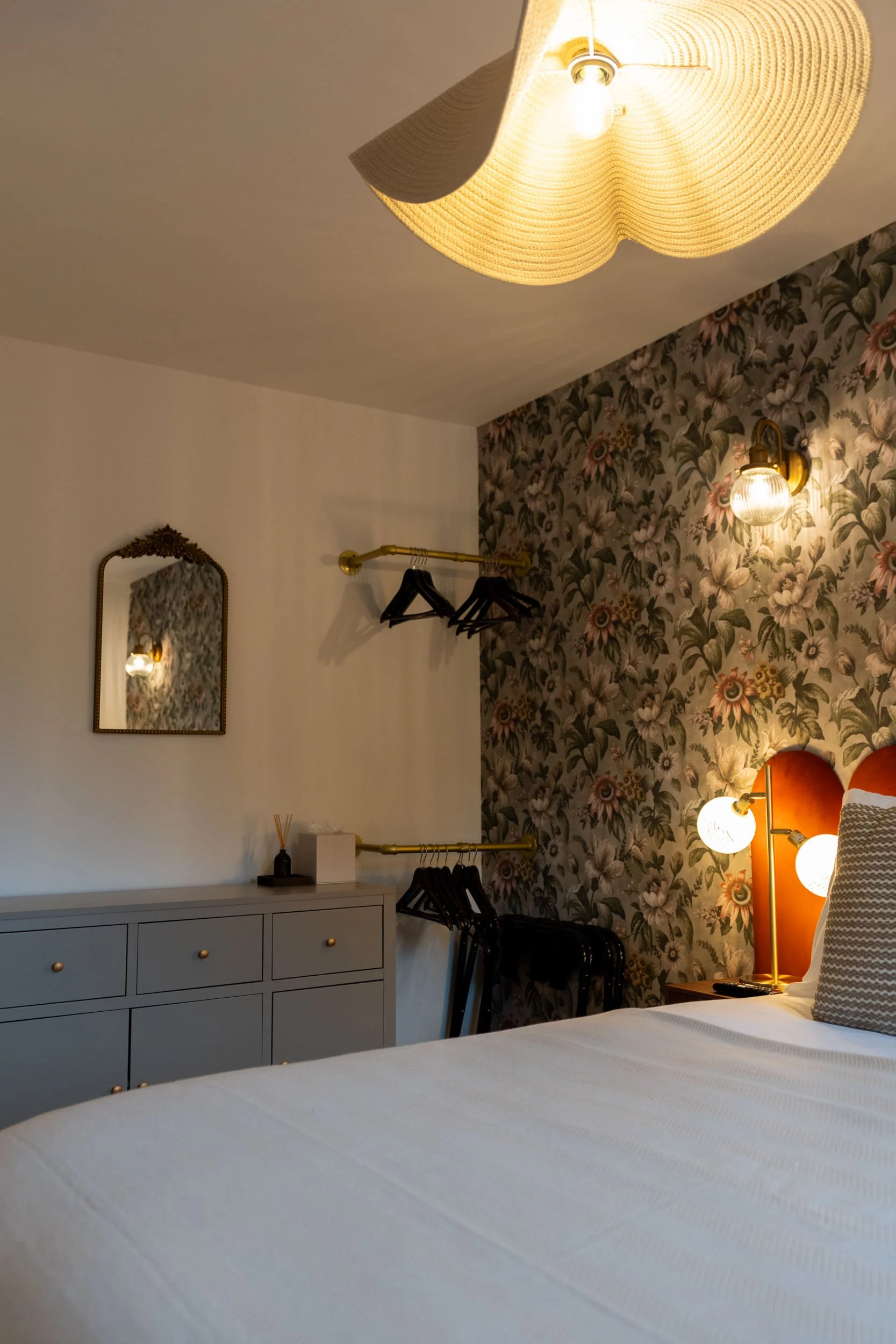 A cozy bedroom featuring a white bedspread, a gray dresser with gold knobs, a decorative mirror, and a floral wallpapered accent wall. There is a gold clothing rack holding black hangers, and warm ambient lighting from a ceiling lamp and wall sconce.