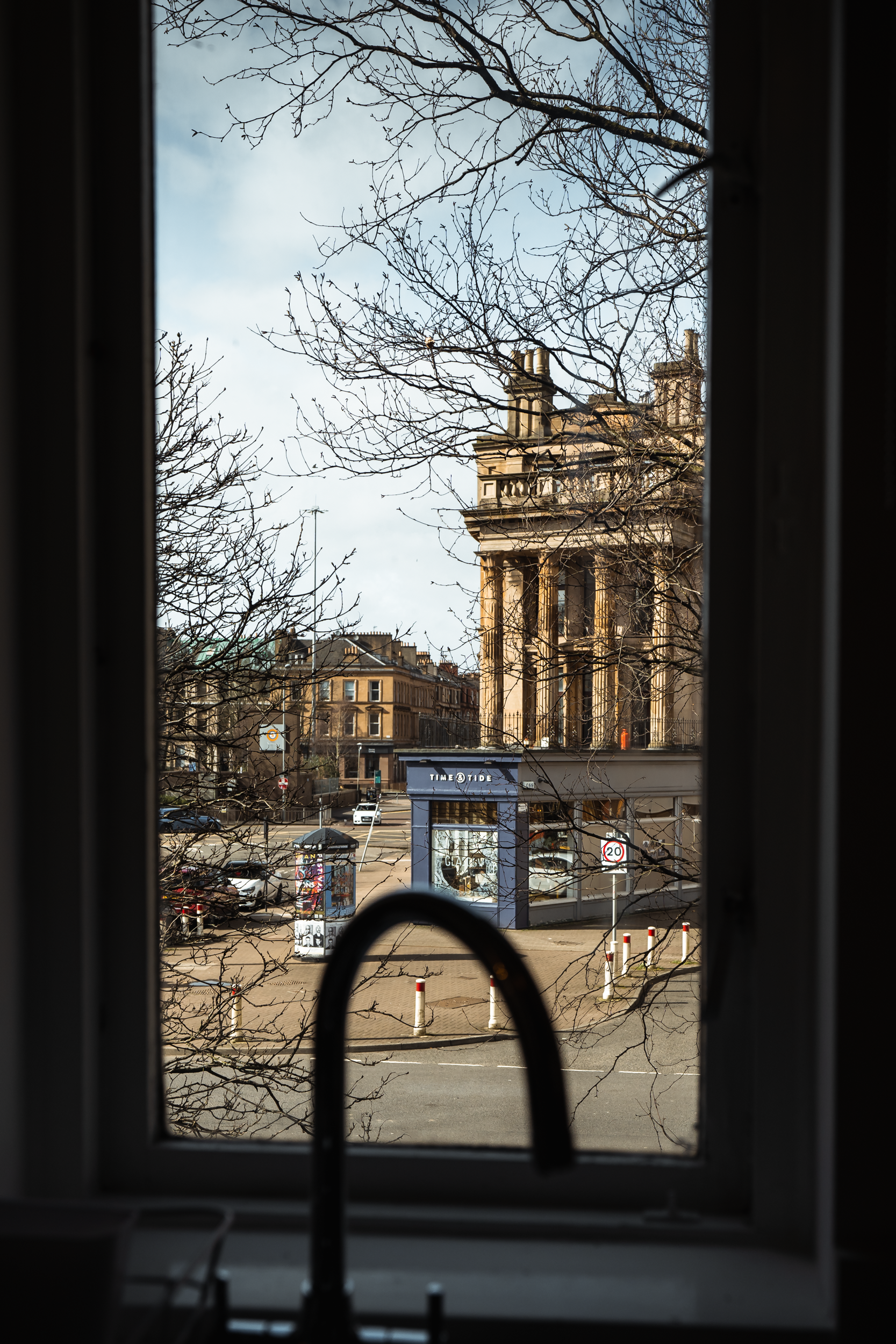 View of a historic building through a window with leafless tree branches in the foreground, and a city street with parked cars and a small kiosk in the background.