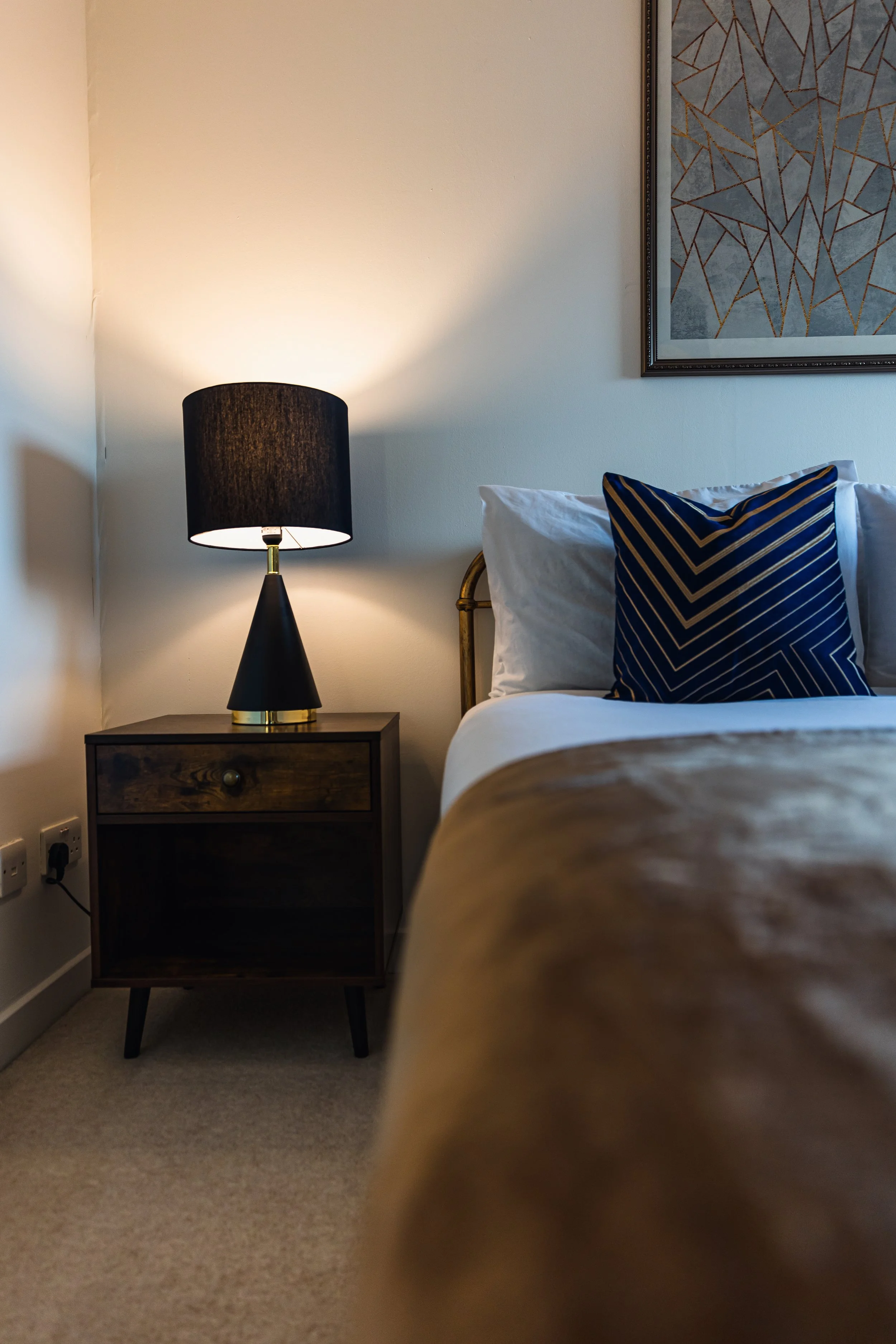 A bedroom corner with a bedside table, a black table lamp, a bed with white bedding and a navy blue pillow with gold lines, and a geometric artwork on the wall.