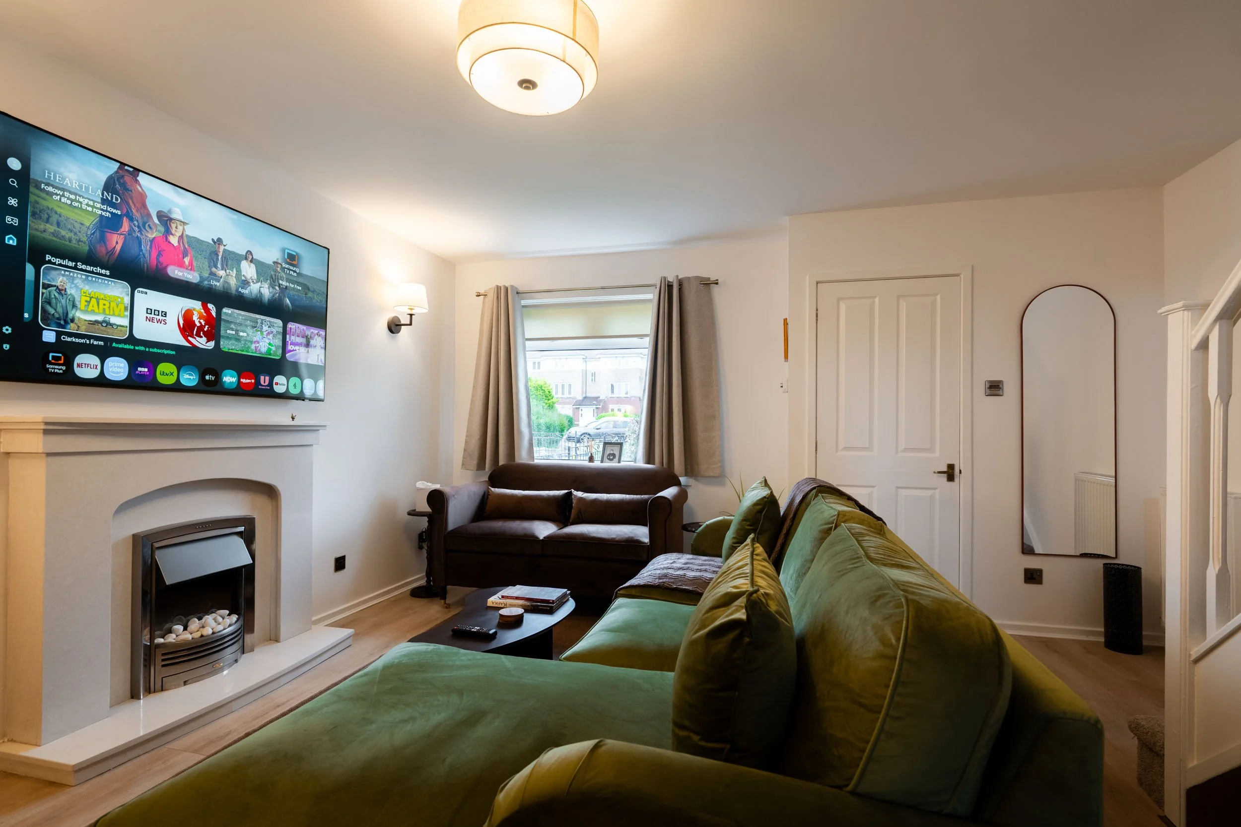 Living room with a mounted flat screen TV, a fireplace with decorative stones, a brown armchair, a green velvet sofa with matching pillows, a coffee table with books and a remote, a window with curtains, a mirror on the wall, ceiling light fixture, a
