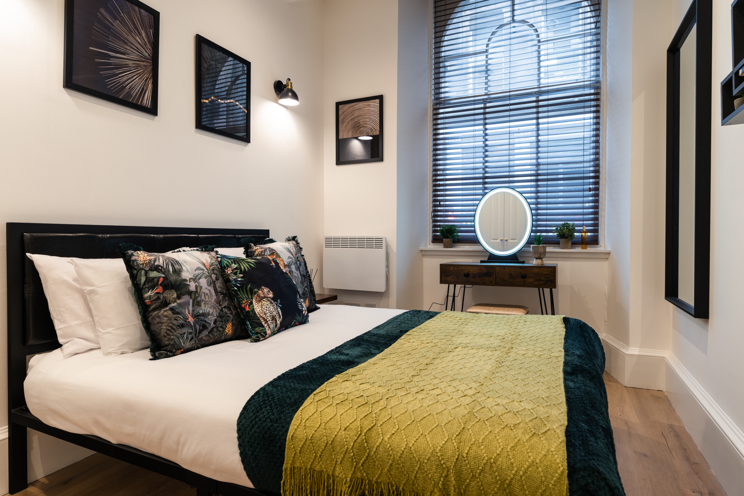 Modern bedroom with bed having tropical-themed pillows, a yellow and green blanket, wall art, a window with blinds, a round illuminated mirror, and small plants on a desk.