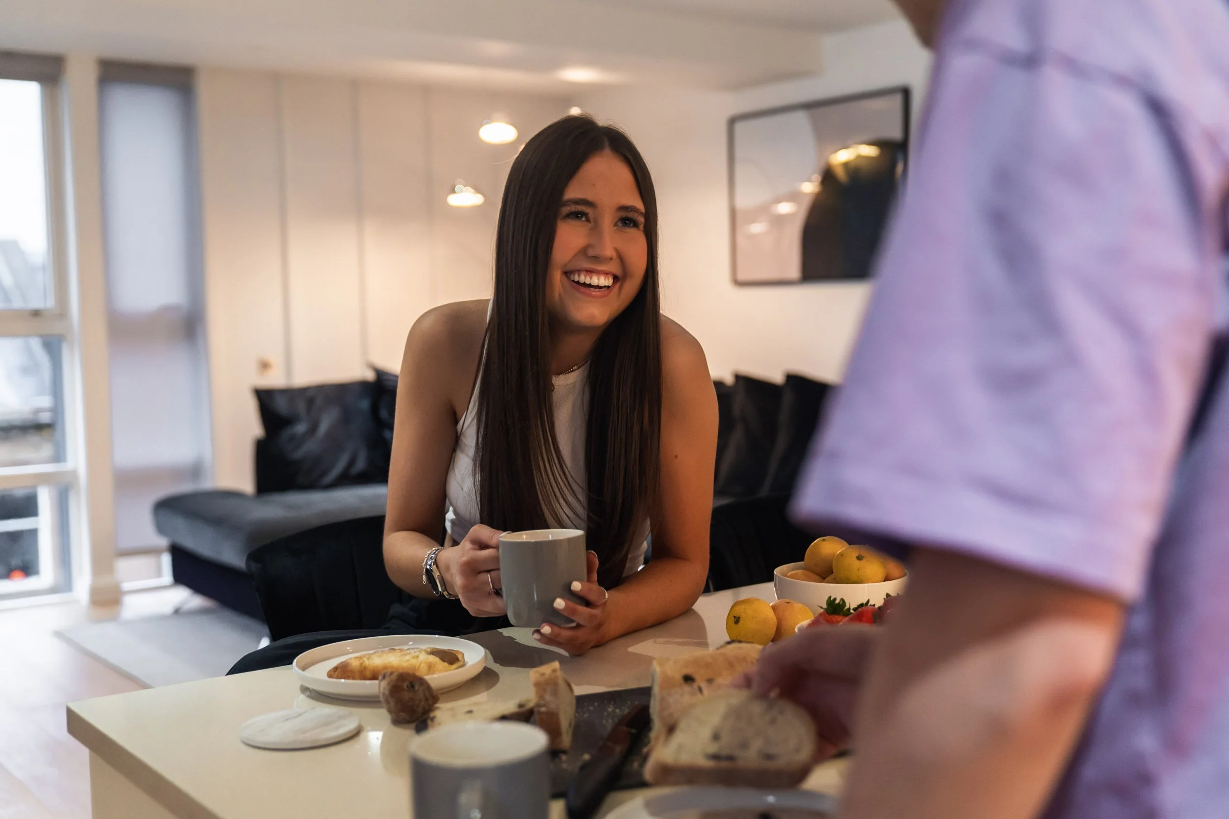 A young woman with long brown hair smiling and holding a coffee mug at a kitchen table with breakfast foods, while a person in a pink shirt serves bread.