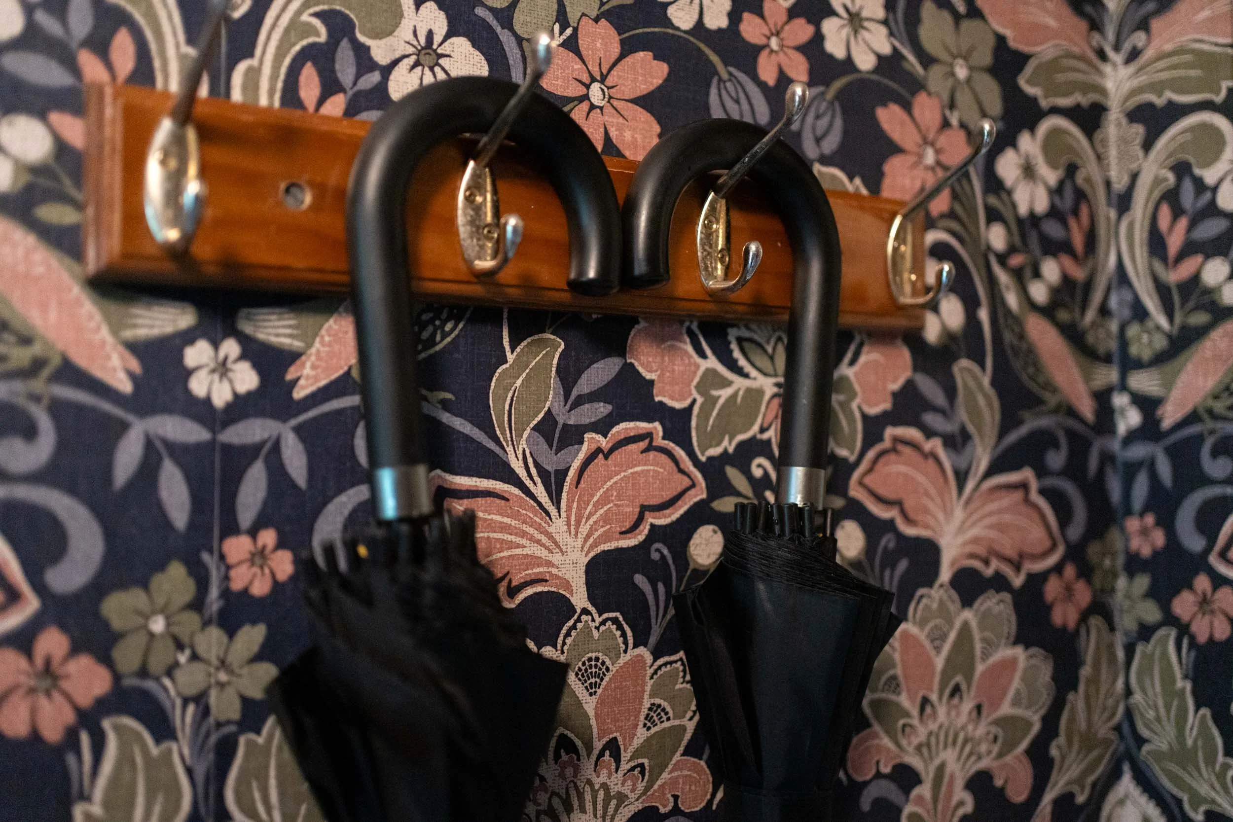 Wooden wall hook rack with silver hooks holding black umbrellas against a floral wallpapered wall.