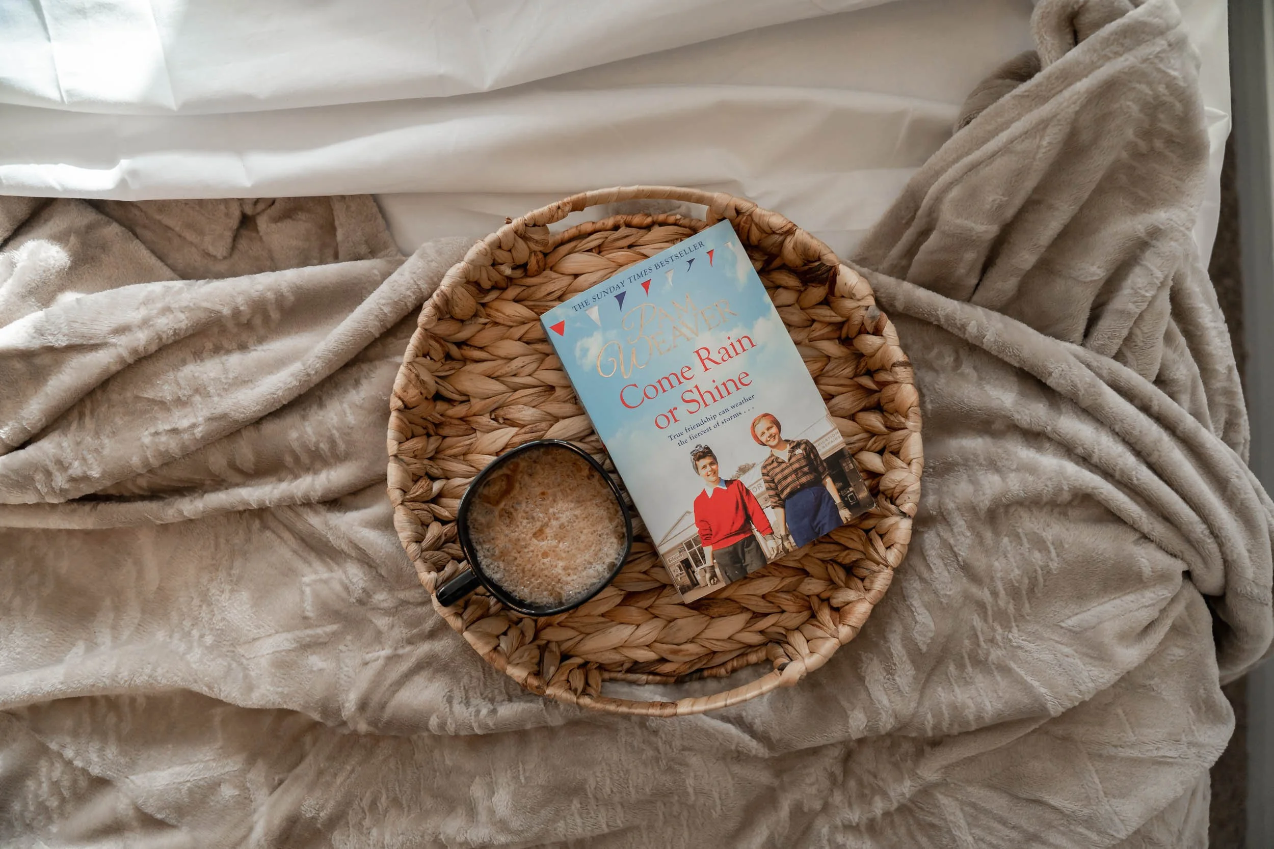 A woven basket containing a book titled 'Come Rain or Shine' by Jan Quaker and a black mug filled with a hot beverage, placed on a beige, fluffy blanket.