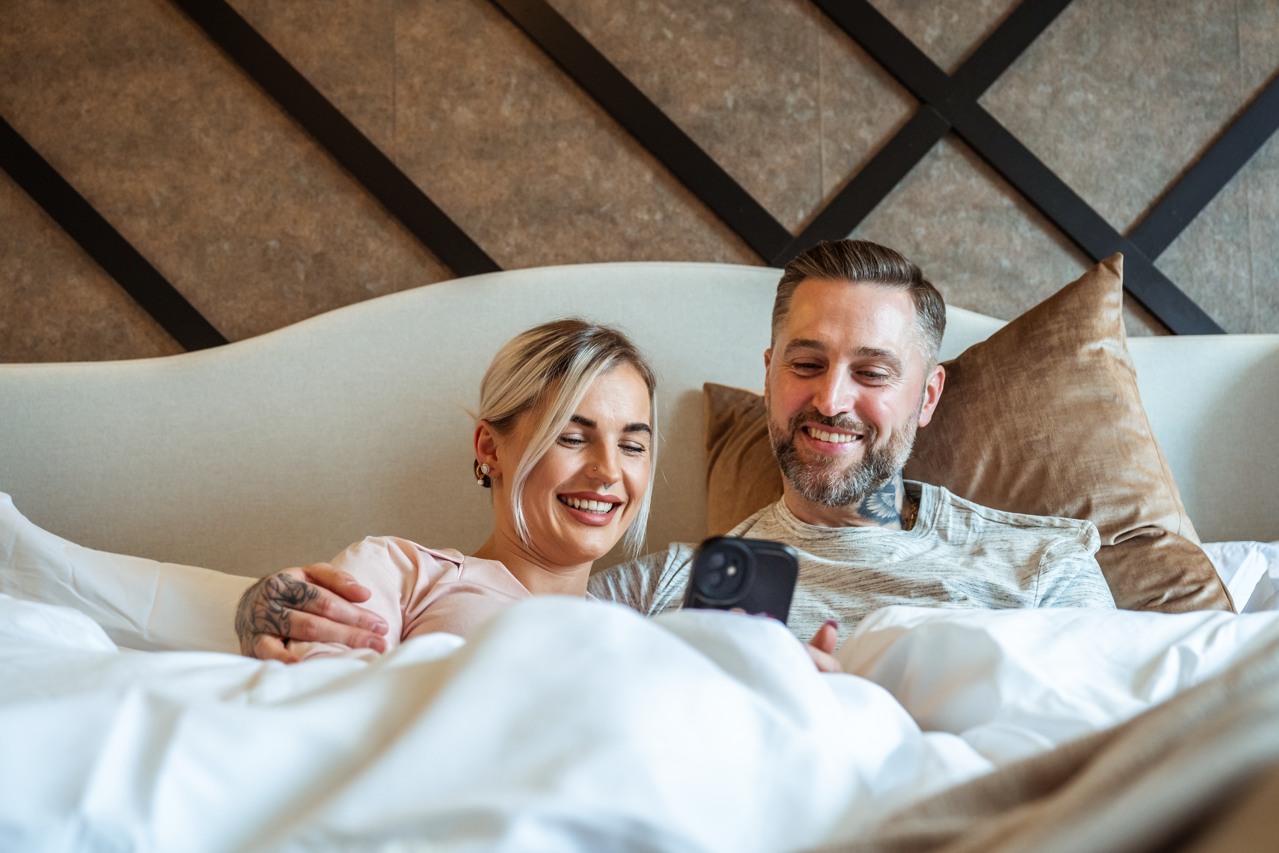 A smiling couple sitting in bed looking at a phone together