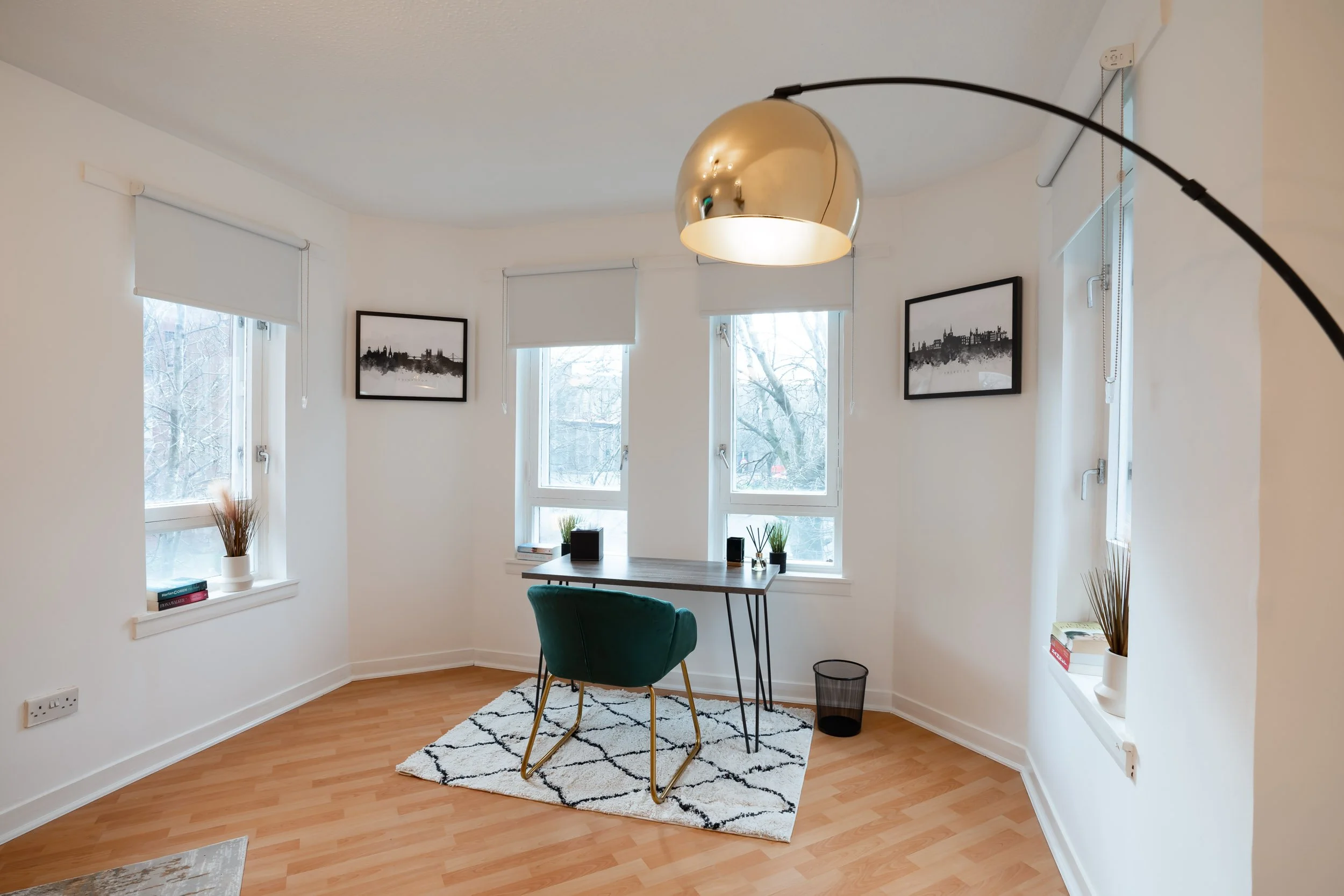 A small home office with three windows, a wooden desk, a green chair, a black trash can, decorative books and plants, black and white cityscape wall art, and a large gold floor lamp.