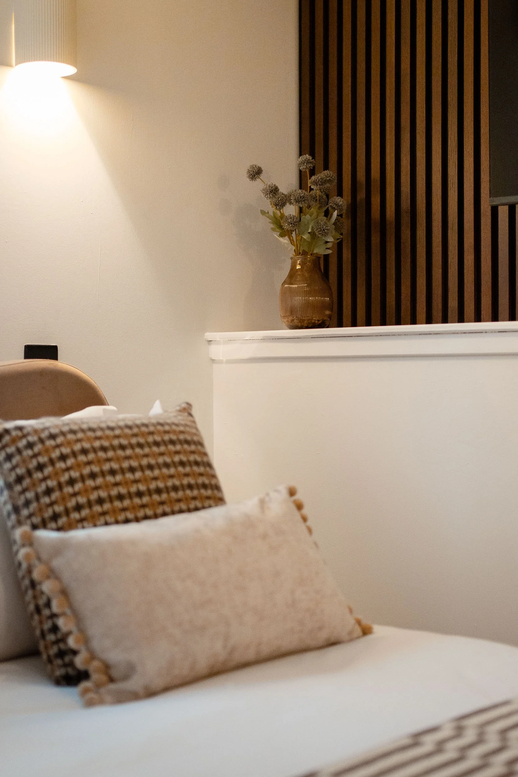 Close-up of beige and patterned pillows on a sofa in a cozy, well-lit living room with a white wall and a wooden wall panel with a vase of flowers.