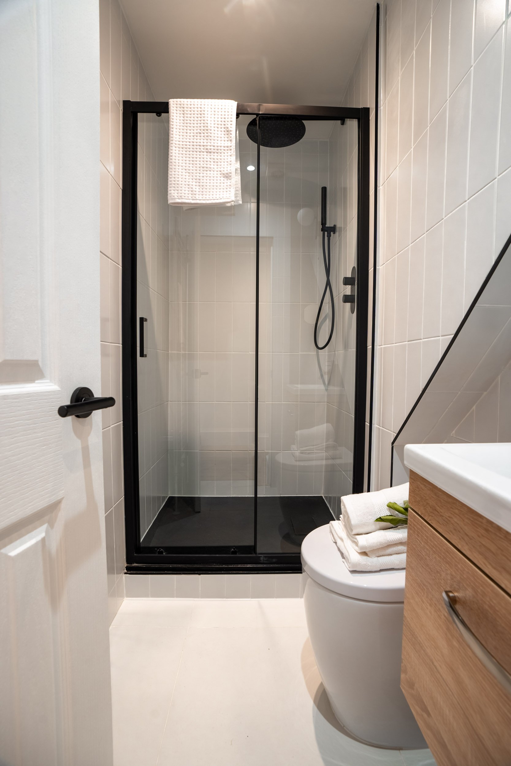 Modern bathroom with a glass-enclosed shower, white tiles, a toilet with folded towels on top, and a wooden vanity.