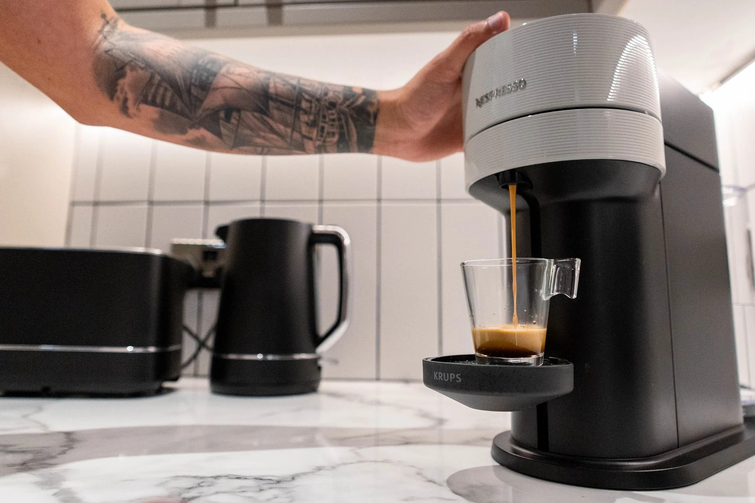 A person with a tattooed arm is pouring espresso from a Nespresso machine into a clear glass cup on a KRUPS coffee scale, with a black kettle and toaster in the background on a marble countertop in a modern kitchen.