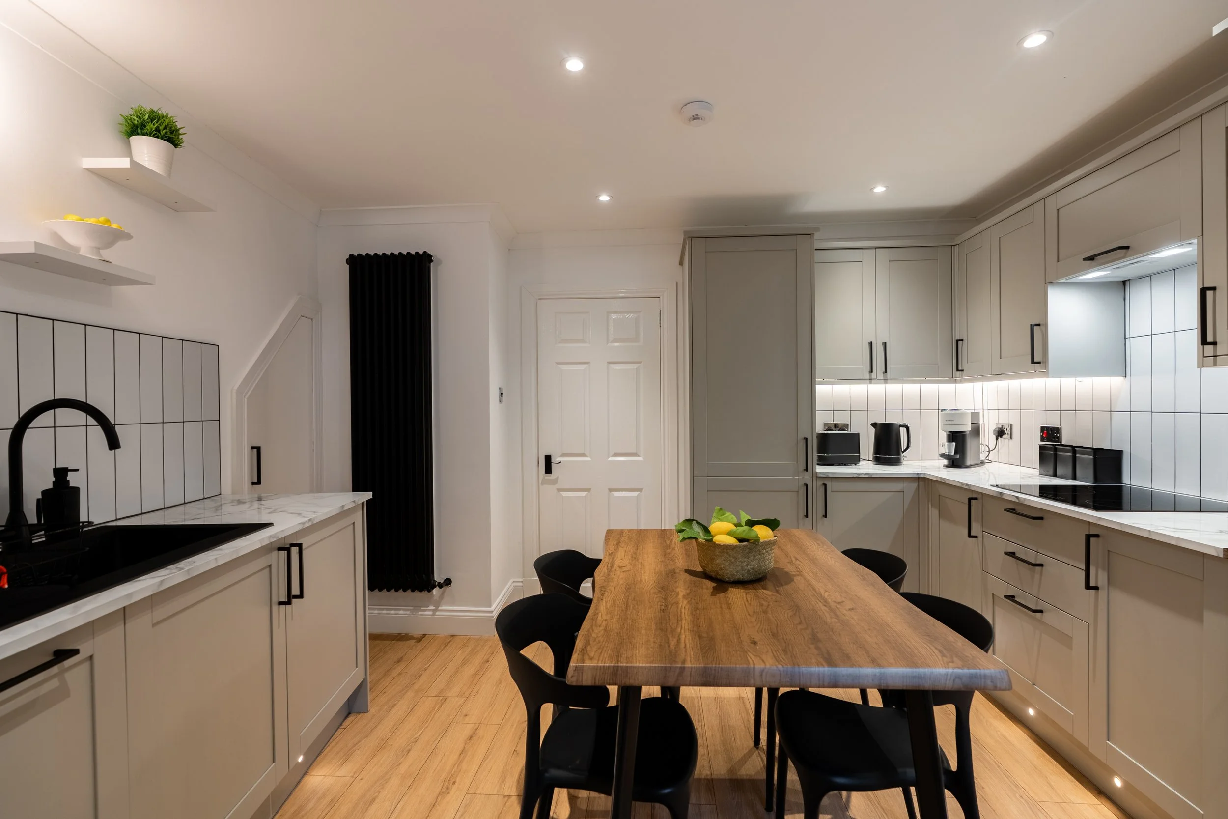 Modern kitchen with white cabinetry, marble countertops, black hardware, and a wooden dining table with black chairs, decorated with a bowl of lemons and green leaves.