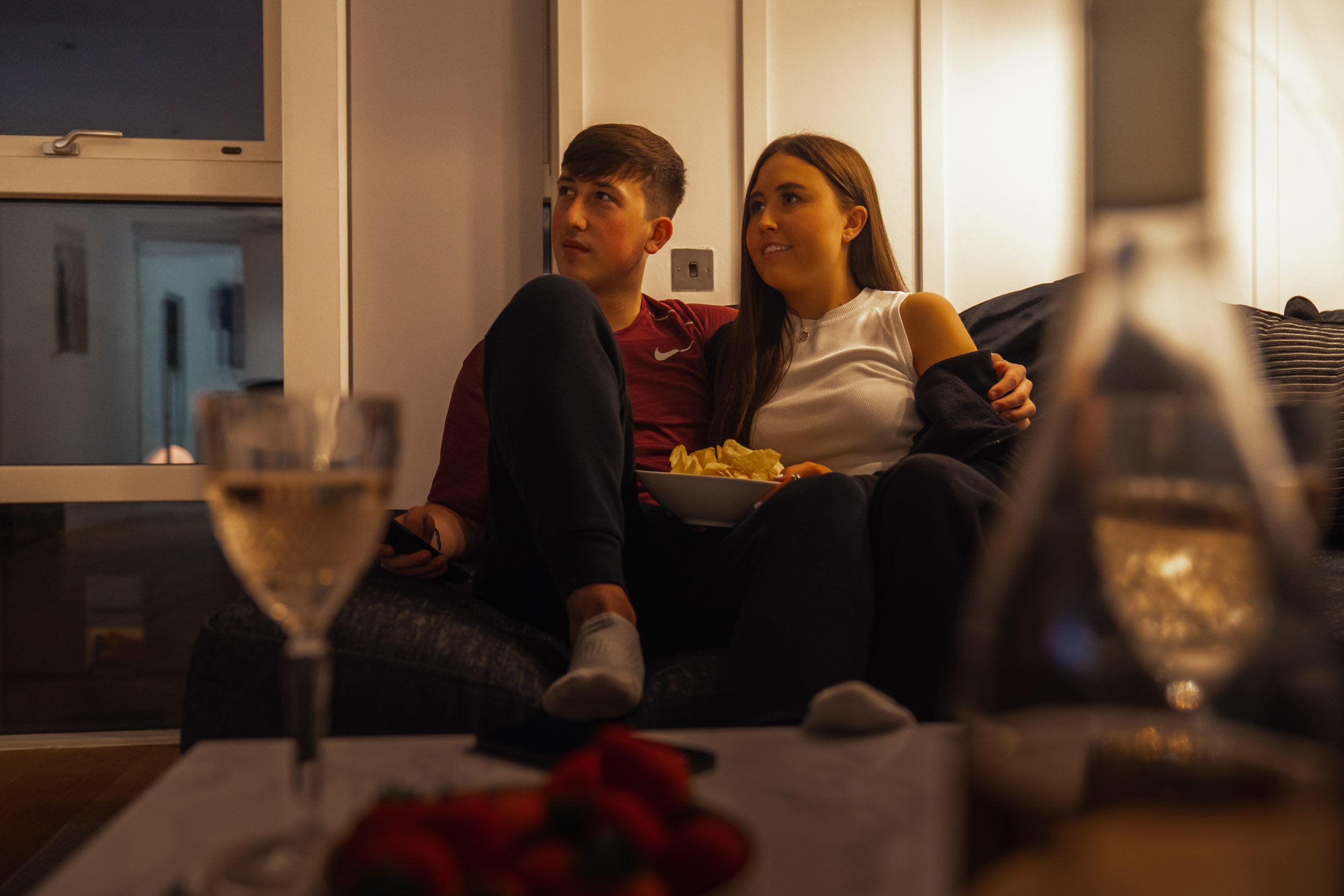 Two young people sitting on a couch in a living room, with snacks, drinks, and a candle on the table in front of them, relaxing together.