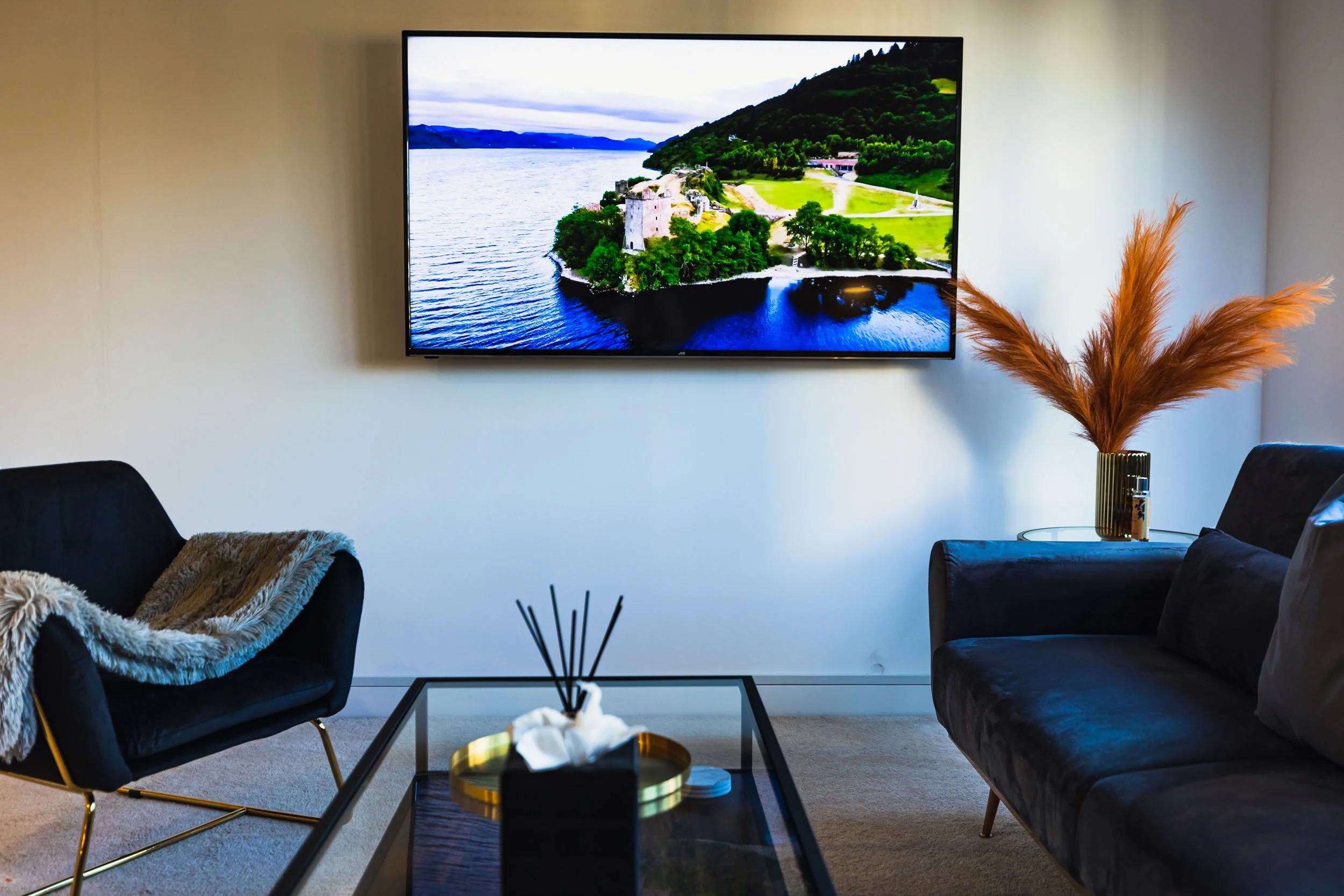 Modern living room with a wall-mounted TV displaying a landscape of a lake and green hills, a black armchair with a faux fur throw, a dark sofa, a glass coffee table with a reed diffuser and tissue box, and a side table with a vase of tall, orange pa