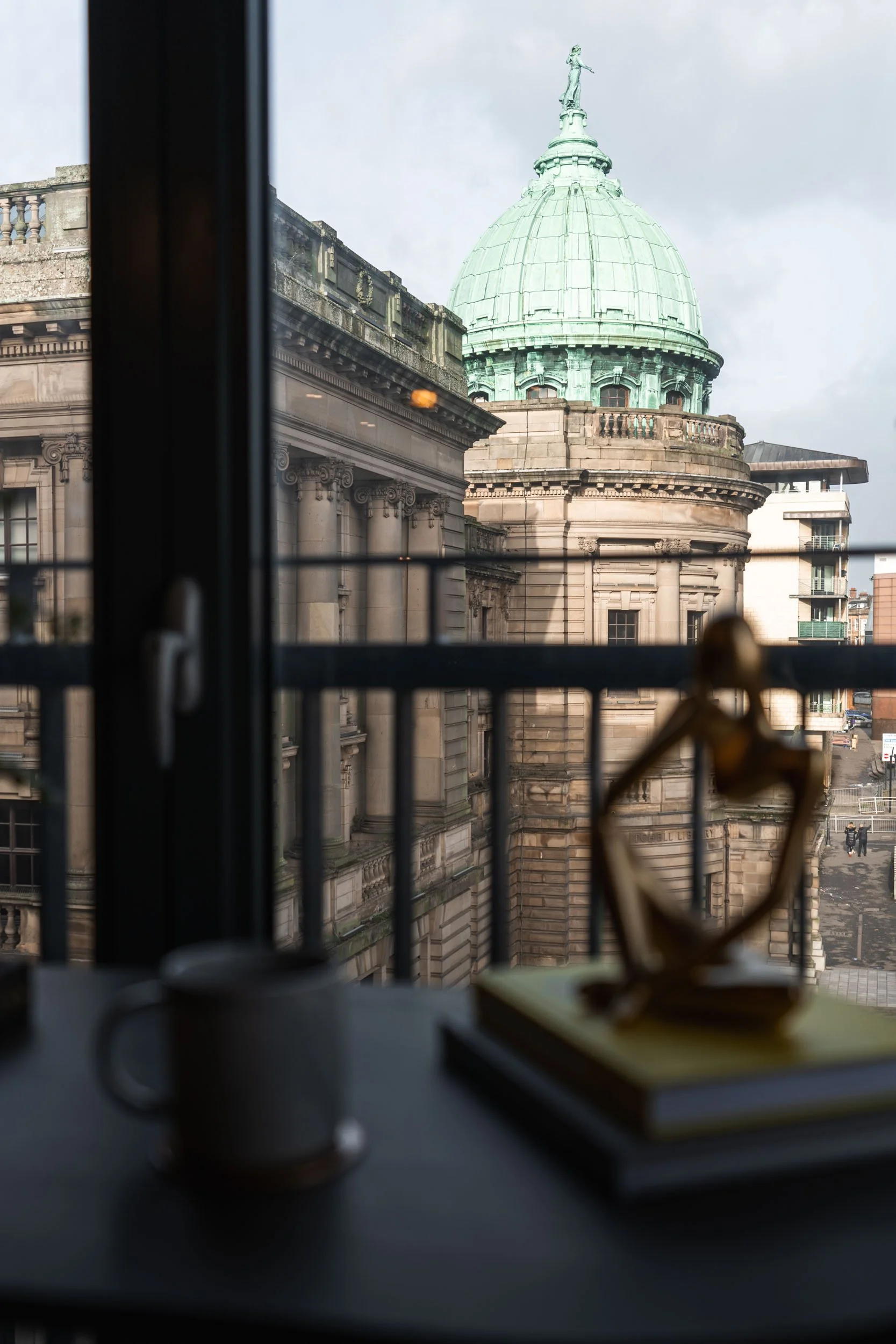 View from inside a room showing a historic building's dome and architecture through a window with a blurred sculpture, books, and a mug in the foreground.