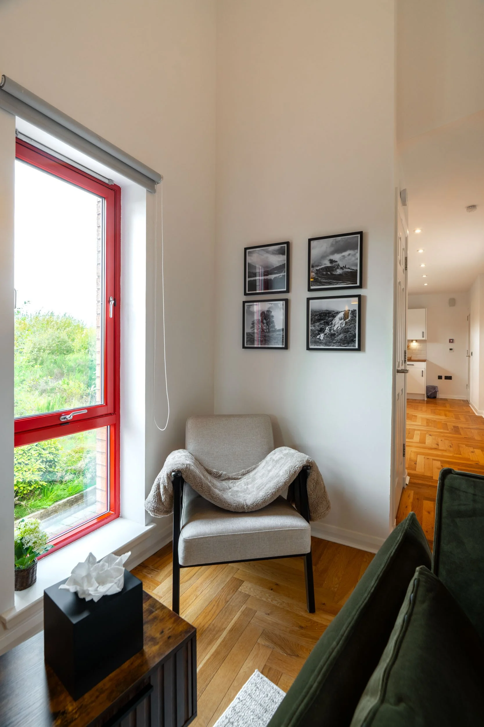 Interior view of a living room with a beige armchair, black and white framed landscape photos on the wall, a side table with a tissue box, a window with a red frame, and a glimpse of a kitchen with wooden floors in the background.
