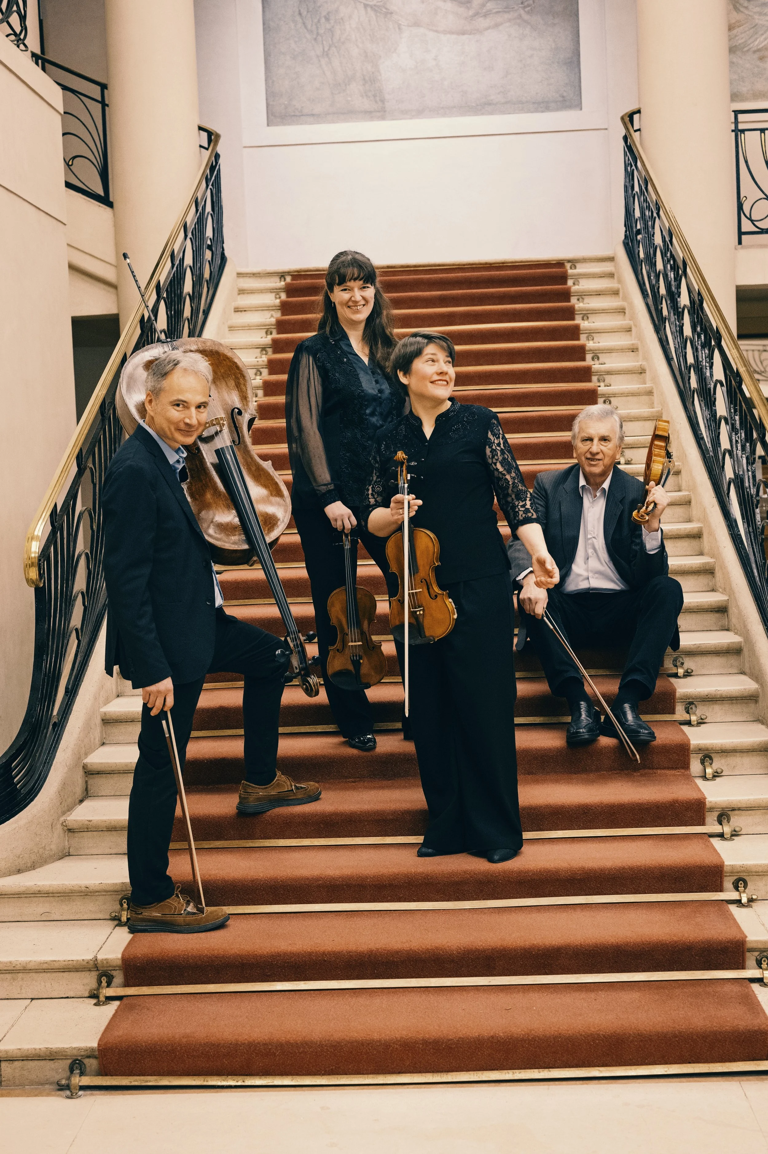 A group of five musicians holding string instruments, standing and sitting on a grand staircase in a classical building, smiling for the camera.