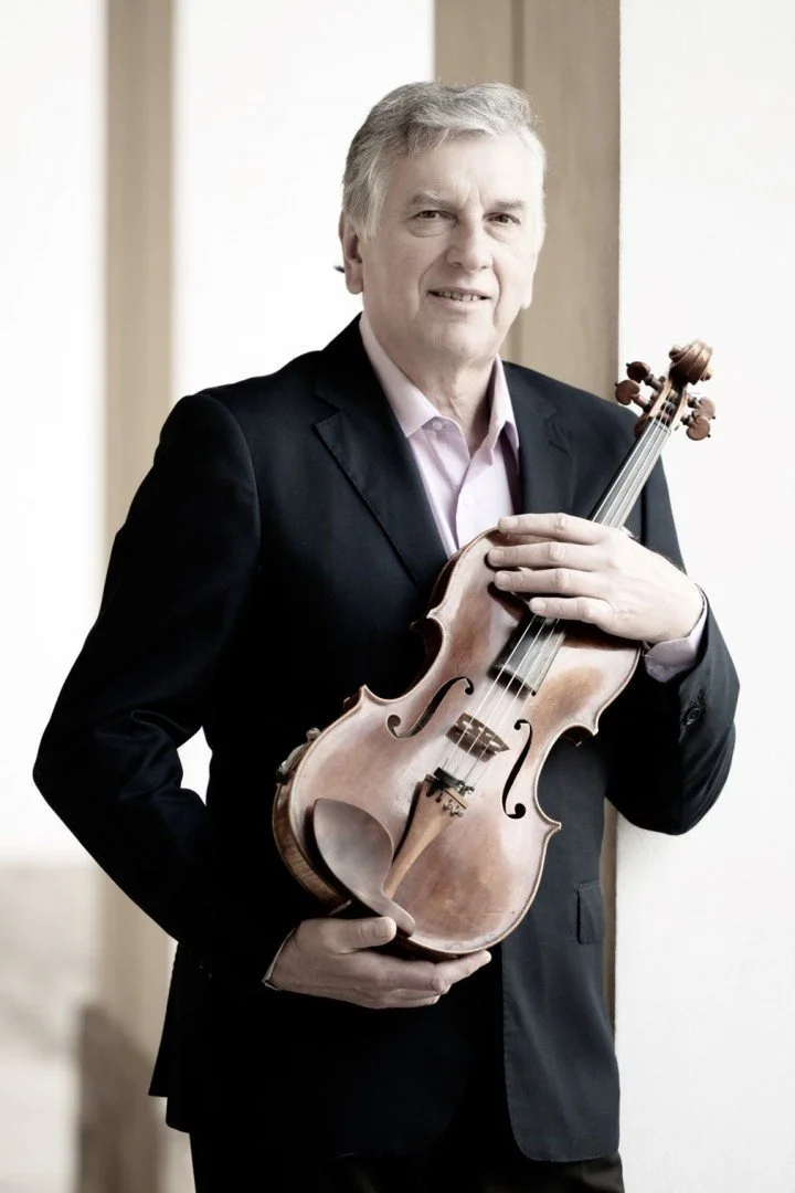An older man in a black suit holding a violin, standing indoors next to a wooden wall.