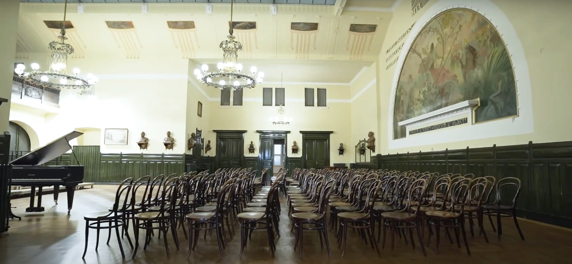 Concert hall with rows of wooden chairs, a black grand piano, and ornate chandeliers in a historic building.