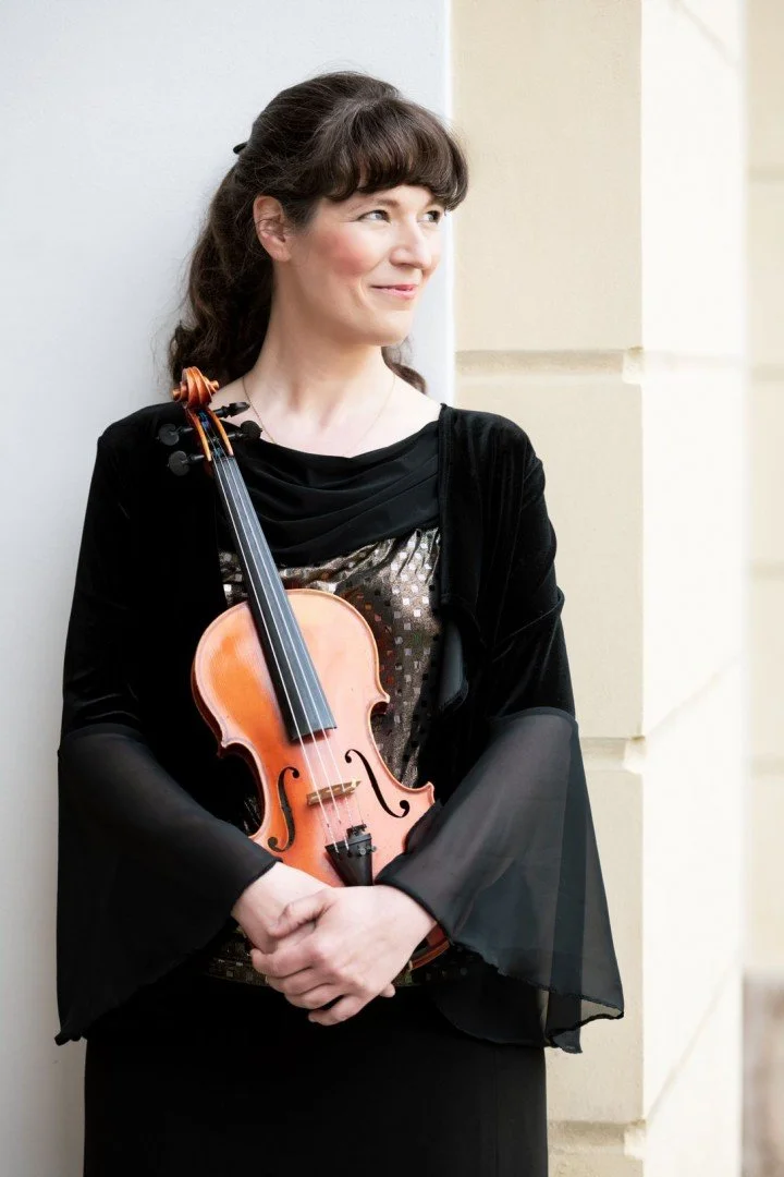 A woman with dark brown hair holding a violin, standing outdoors against a light-colored wall, smiling and looking to her right, dressed in a black outfit with sheer sleeves.