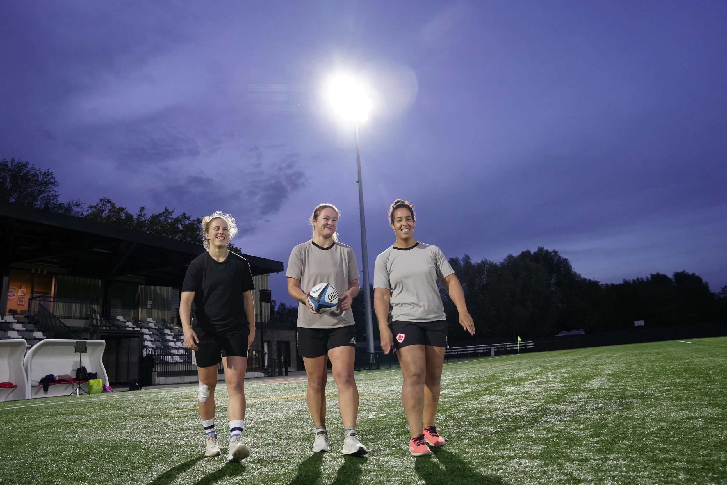 Trois femmes marchant sur un terrain de rugby éclairé par un lampadaire en soirée, avec un ciel nuageux en arrière-plan.
