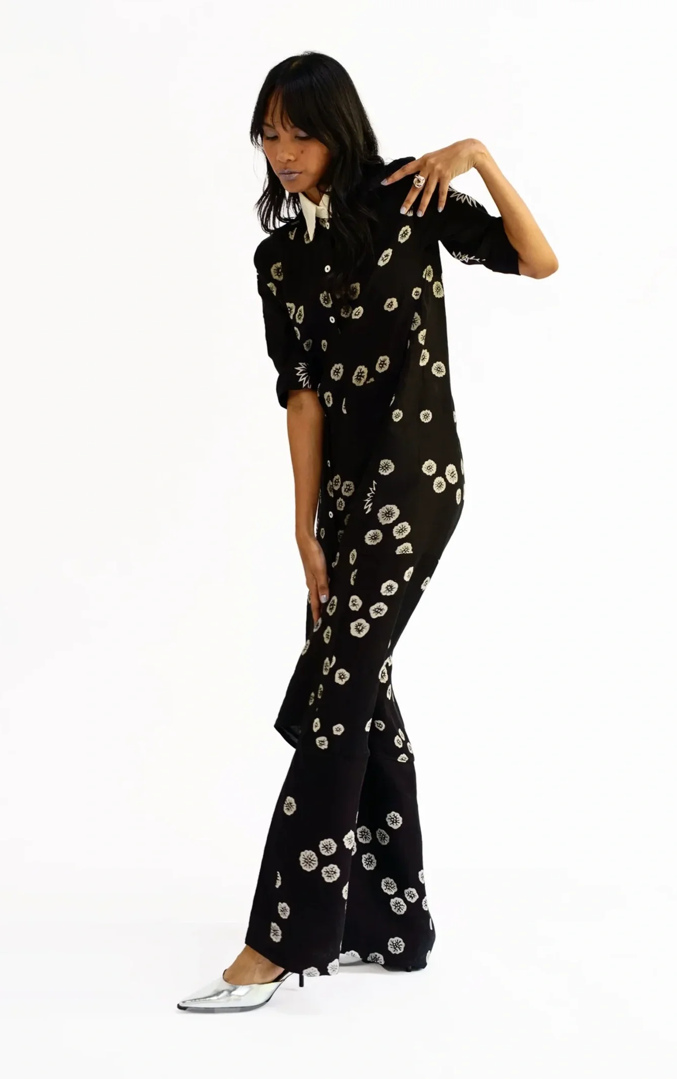 Woman in an oversized black shirtdress hand-painted with a scattered cream floral dot motif, one hand raised to her shoulder, from Hypatia's Batik Tulis textile design line.