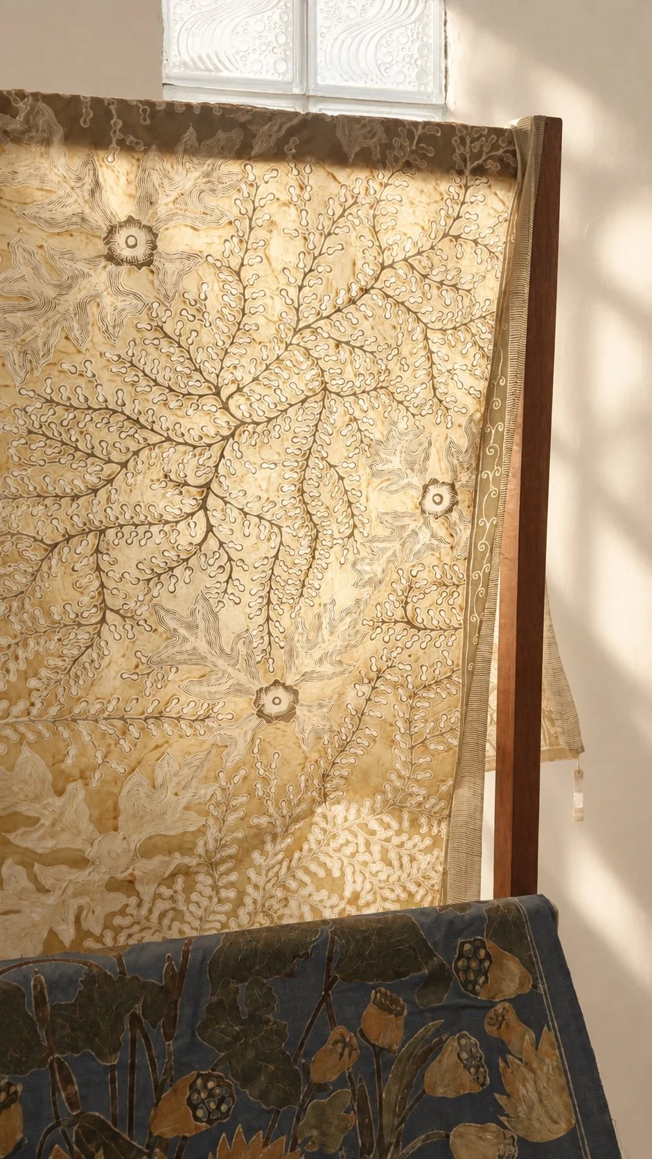 A length of Hypatia Batik Tulis drying over a wooden frame by a window, its golden fern-and-flower pattern glowing in the light, with a blue cloth drawn with lotus pods and leaves folded beneath it.