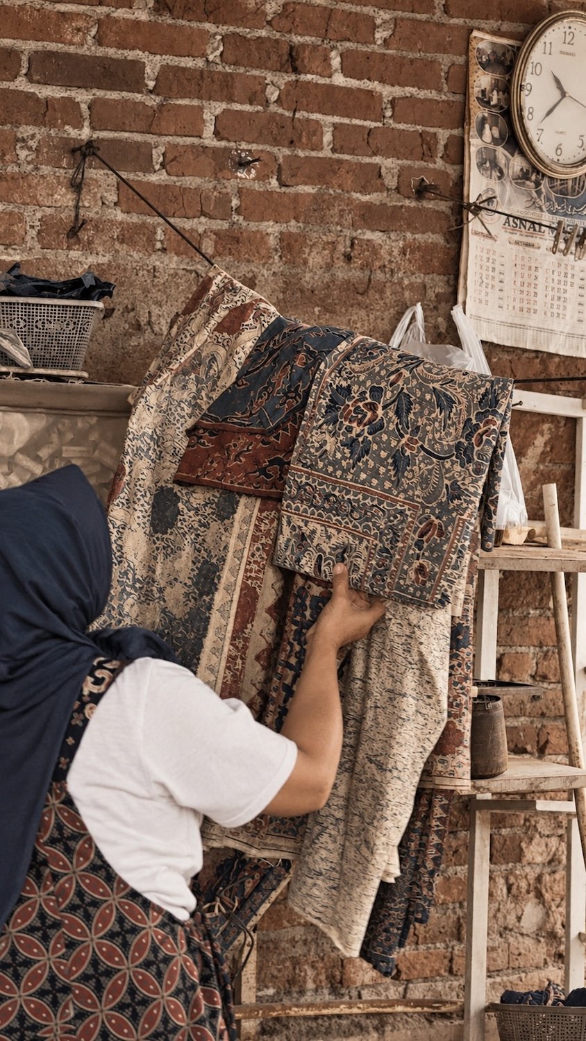 HYPATIA Batik Tulis - artisan inspecting finished cloths against the workshop wall - hand-drawn wax-resist batik in rust, indigo, and cream.