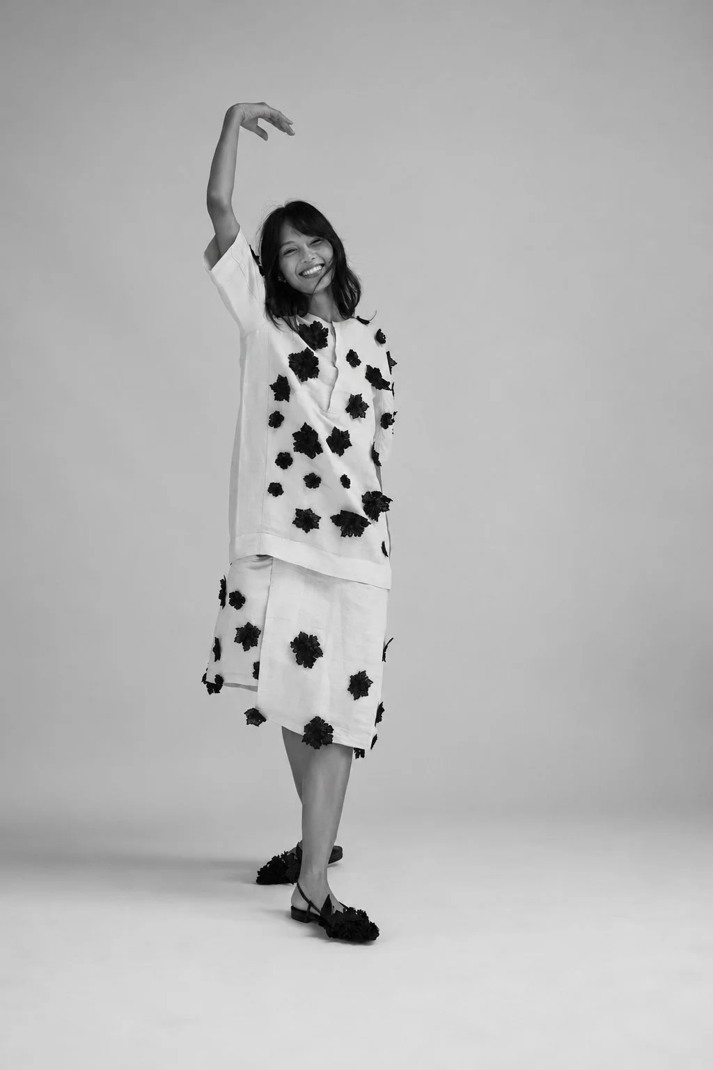HYPATIA Original 17 – Smiling model in a white linen two-piece covered in hand-placed black floral appliqué, one arm raised overhead, caught mid-movement, black flats echoing the appliqué, black-and-white studio portrait