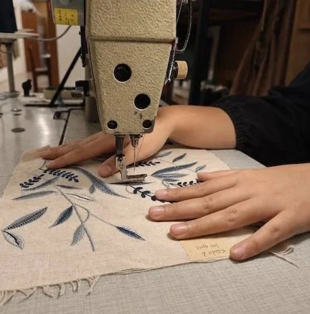 Close-up of the machine's needle and presser foot stitching a densely worked section of the blue and pale-blue leaf-and-vine motif.