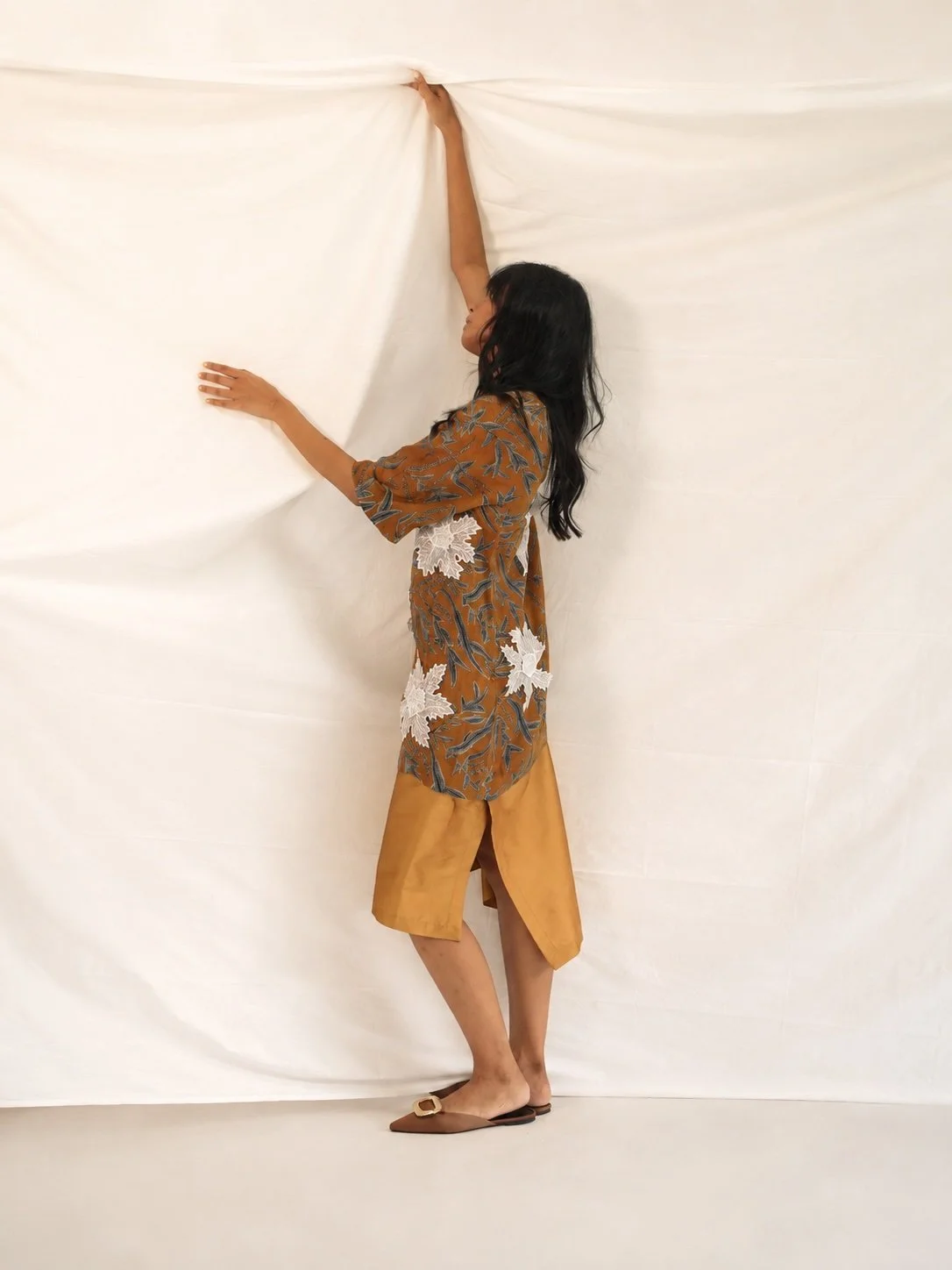 Woman in profile raising a length of raw Hypatia Serat cloth overhead, wearing a rust-brown Batik Tulis dress with Sulam floral appliqué.