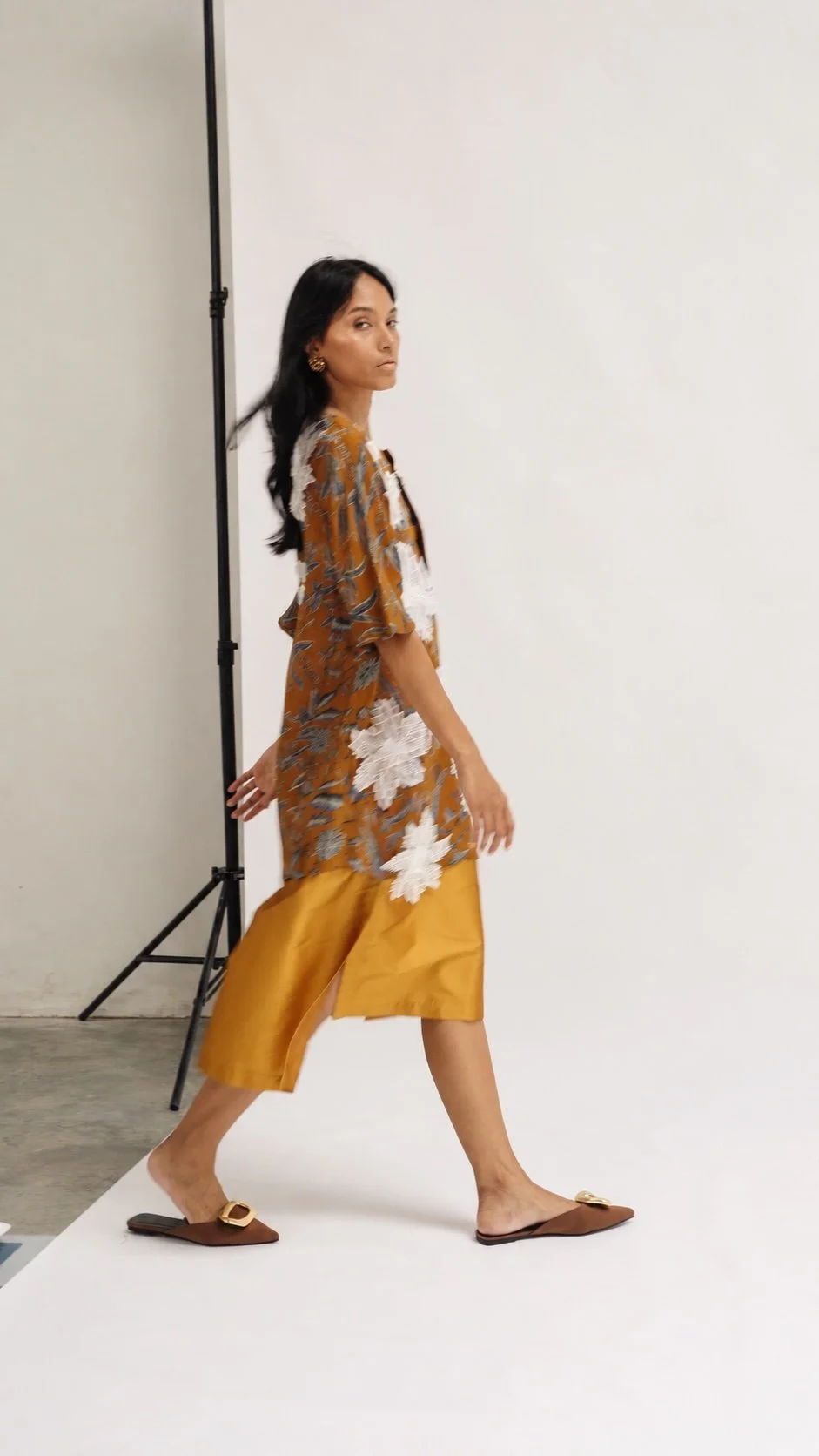 HYPATIA Combinables – woman walking past a studio light stand, glancing over her shoulder, mustard Batik Tulis top with blue-grey leaf pattern and raised white floral appliques, plain golden-mustard skirt with side slit, brown mules